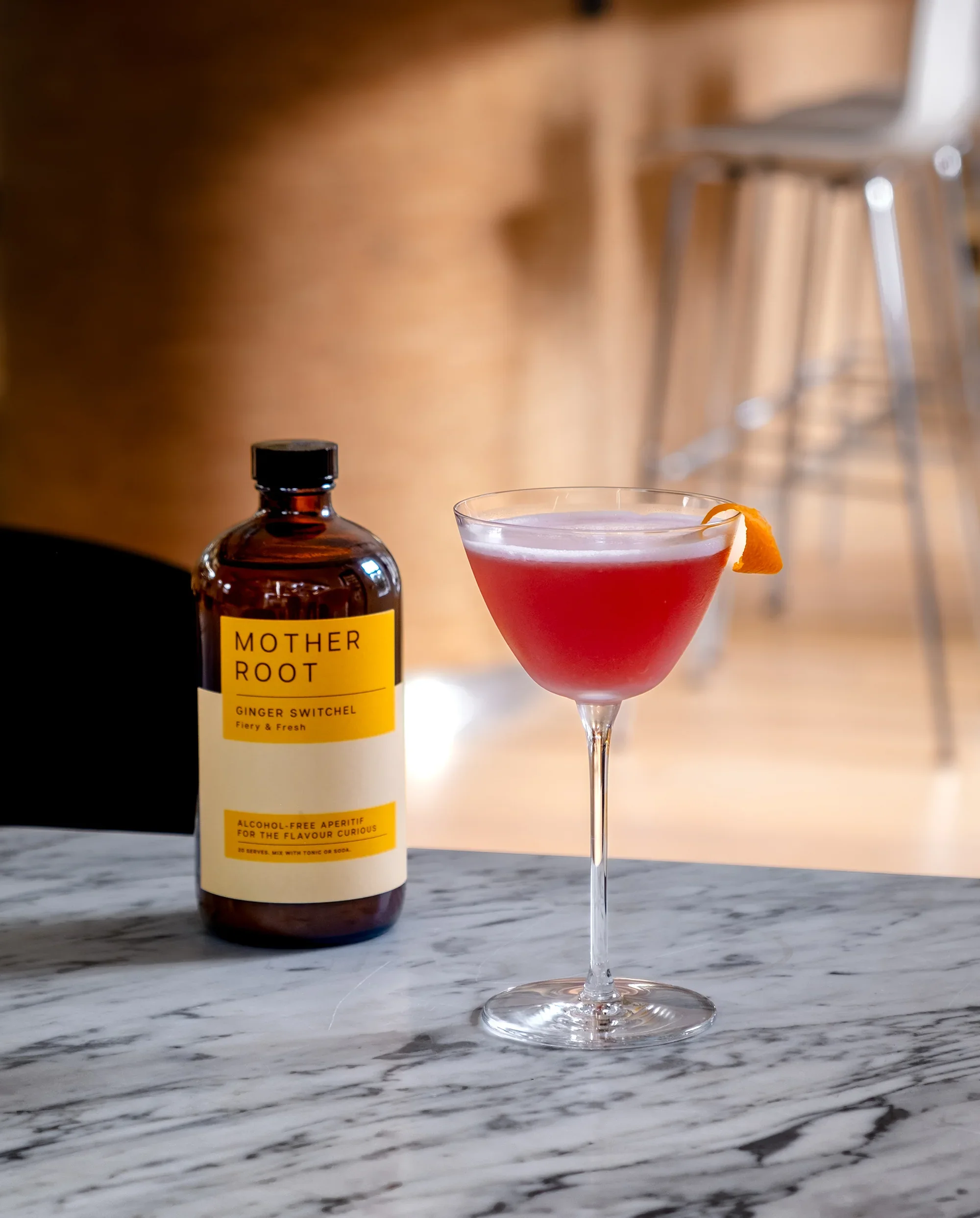 A pink cocktail in a martini glass with an orange twist garnish, a bottle labeled 'Mother Root Ginger Switch' behind it, and a blurred background of bar stools and wooden paneling.