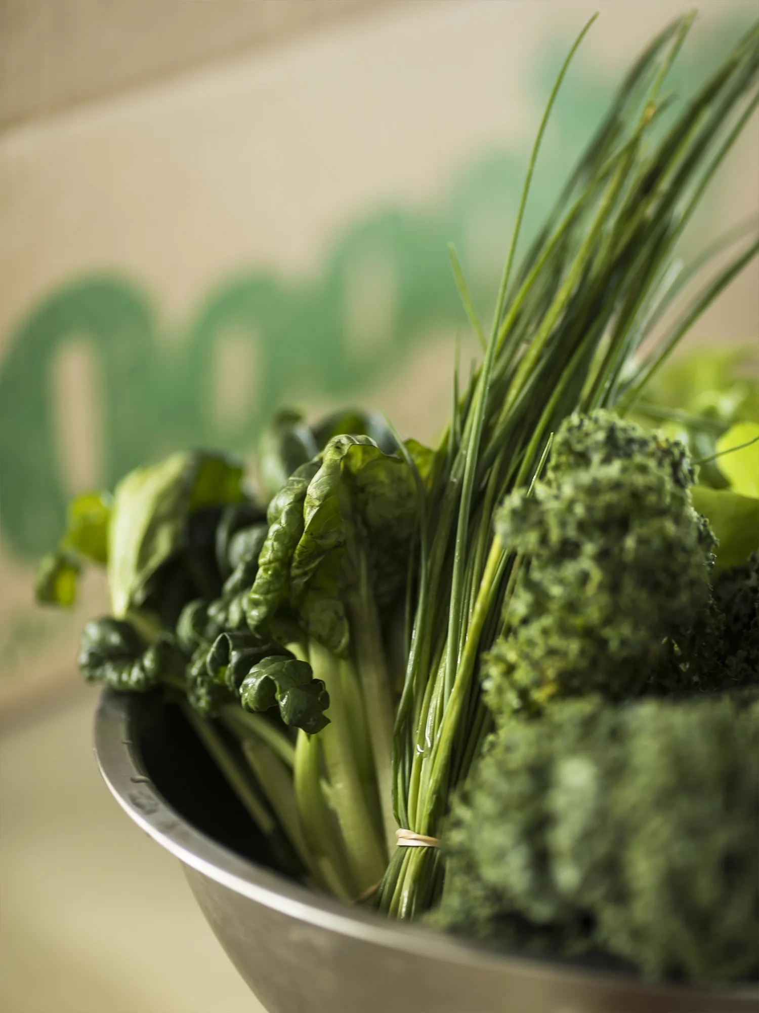 Close-up of fresh green leafy vegetables, including lettuce, broccoli, and chives, in a metal bowl.