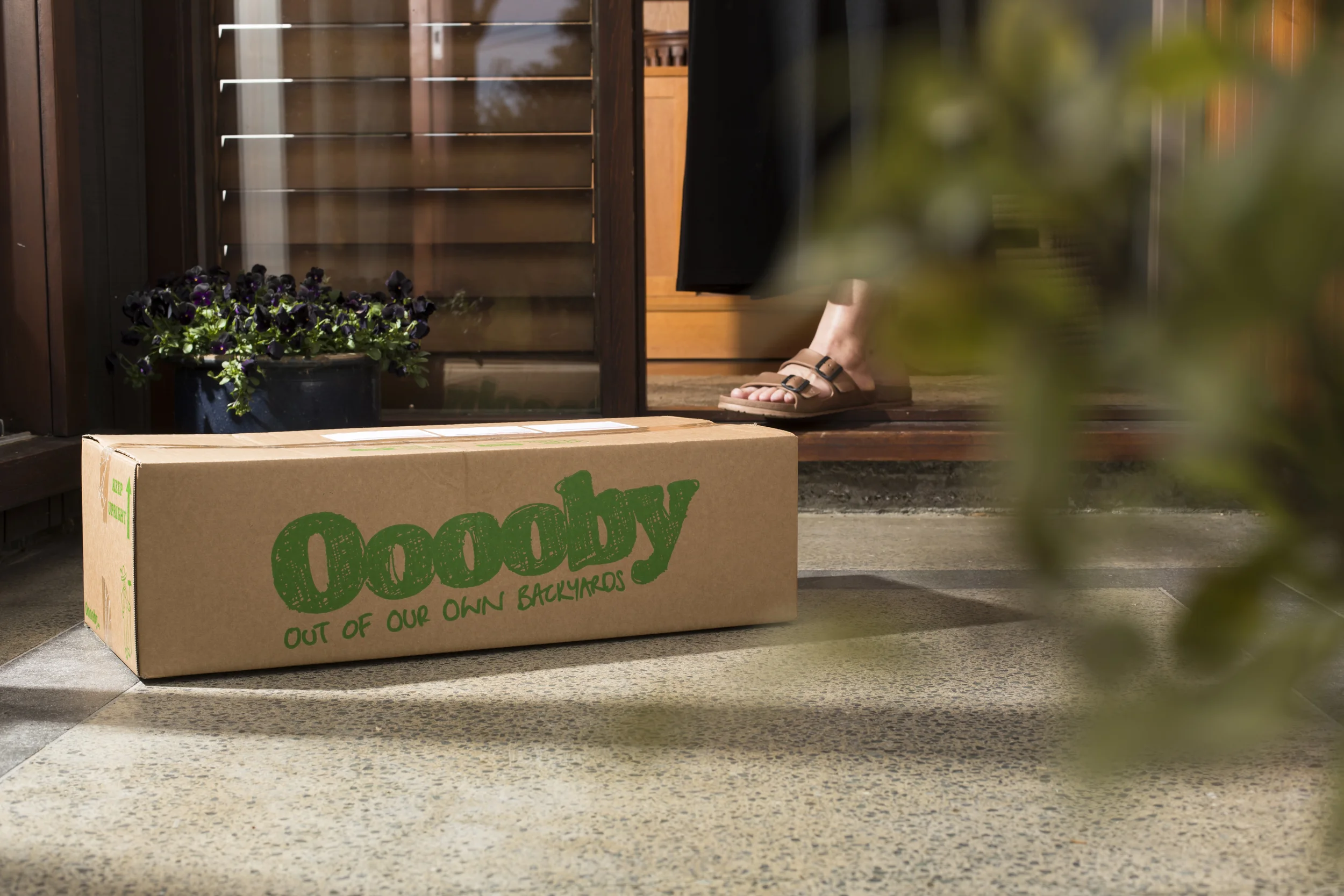 Close-up of a cardboard box labeled 'Community' with the phrase 'out of our own backyards,' placed on a porch in front of a wooden door with glass panels. A pair of sandals is visible on the steps. A potted plant with purple flowers is on the left side.