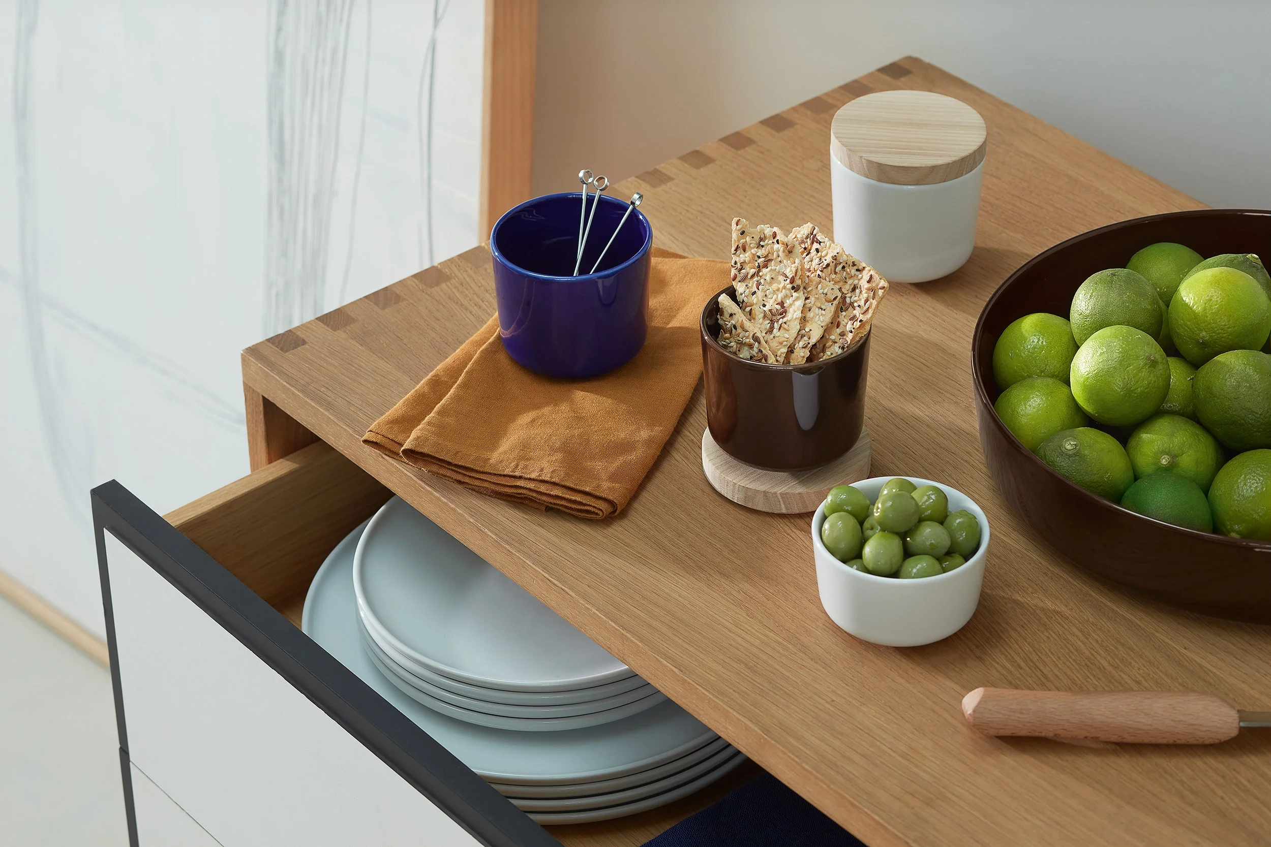 A wooden kitchen island with a bowl of limes, a small bowl of green olives, a dark brown cup of sesame snacks, a blue cup with metal picks inside, and a white container with a wooden lid. There are also some white plates stacked in a drawer below.
