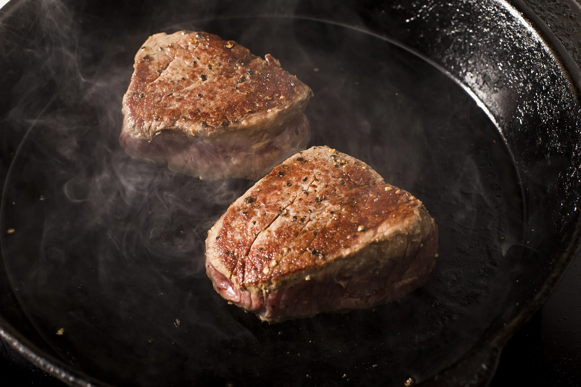 Two seasoned steak chunks cooking in a black cast iron skillet with steam rising.