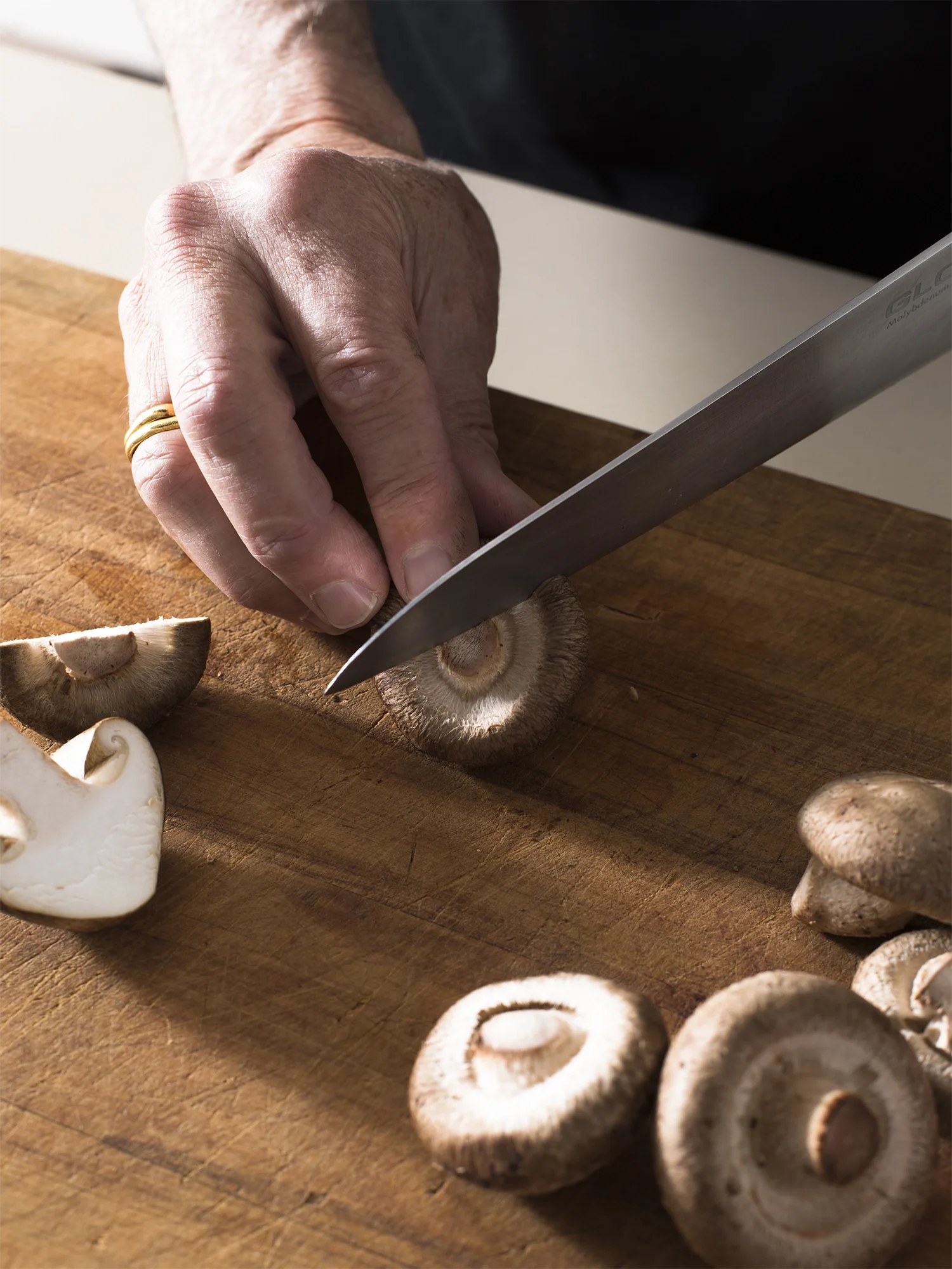A person's hand is slicing a mushroom on a wooden cutting board with a chef's knife.