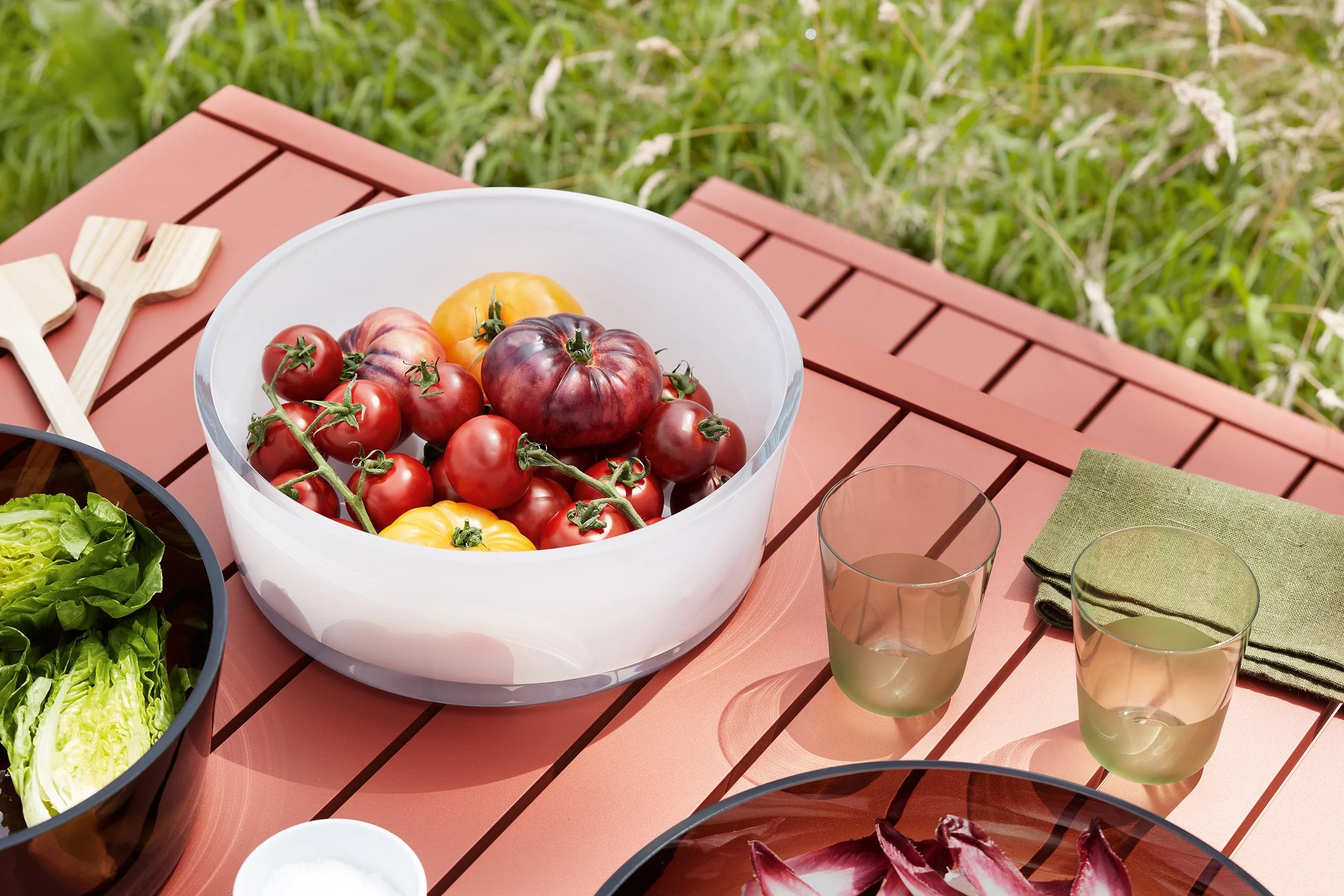 A bowl of fresh heirloom tomatoes on a red wooden outdoor table, with a green grassy background, alongside glasses of drink and a green napkin.
