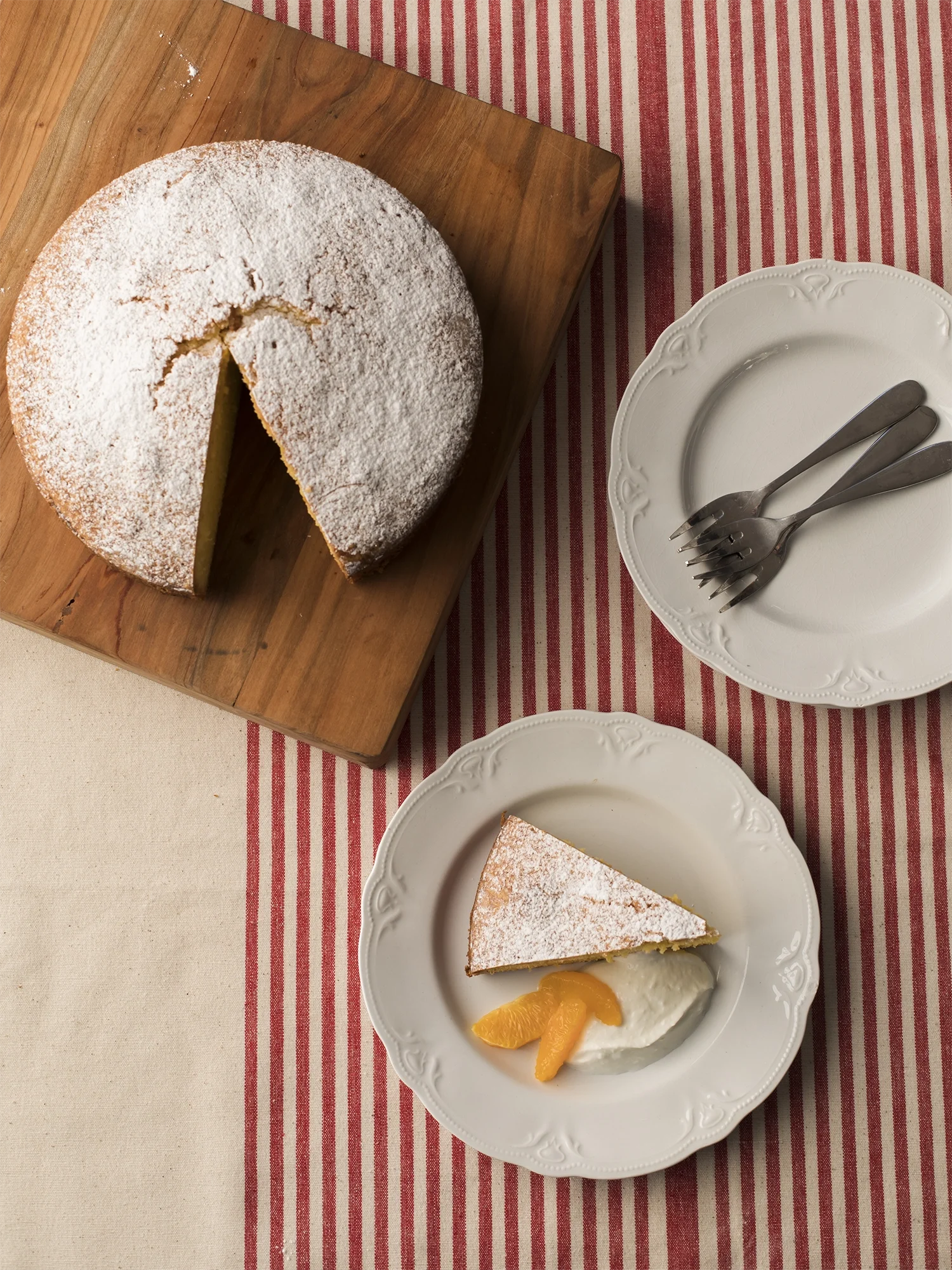 A round powdered sugar cake with a slice removed, placed on a wooden cutting board. A plate with a slice of cake, whipped cream, and orange slices. Two forks are on an empty decorative white plate, on a striped red and white tablecloth.