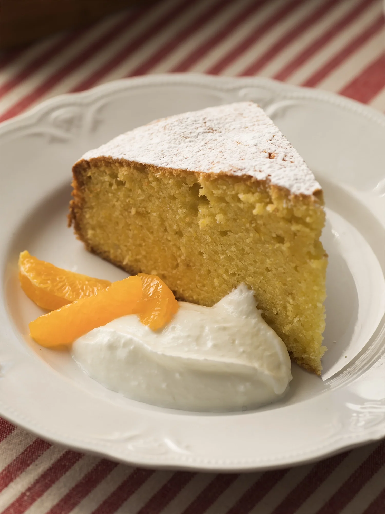 Slice of sponge cake with powdered sugar on top, served with whipped cream and orange slices on a white plate.