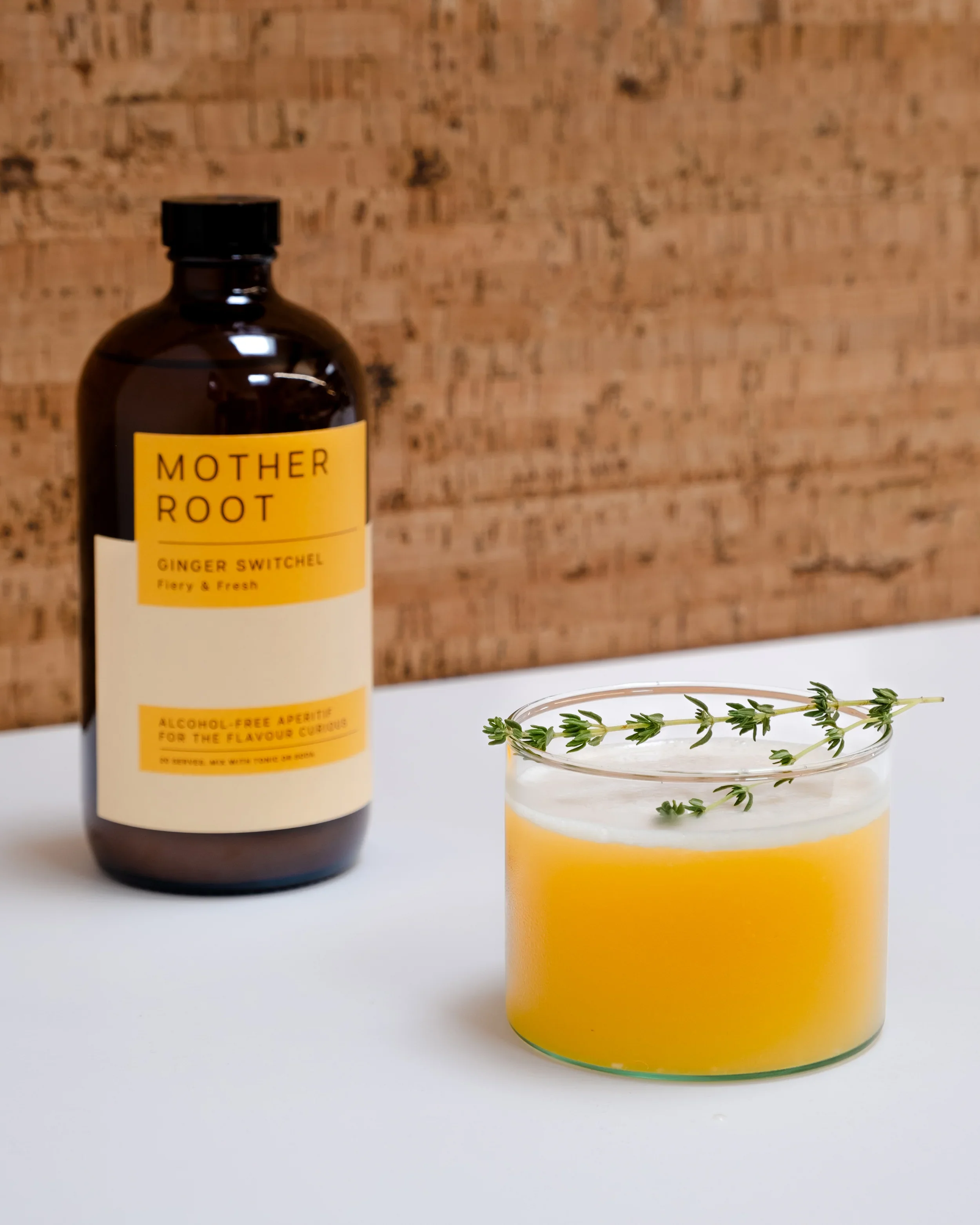 A bottle labeled 'Mother Root Ginger Switchel' with a glass containing a yellow liquid and garnished with fresh thyme sprigs, set against a wooden background.