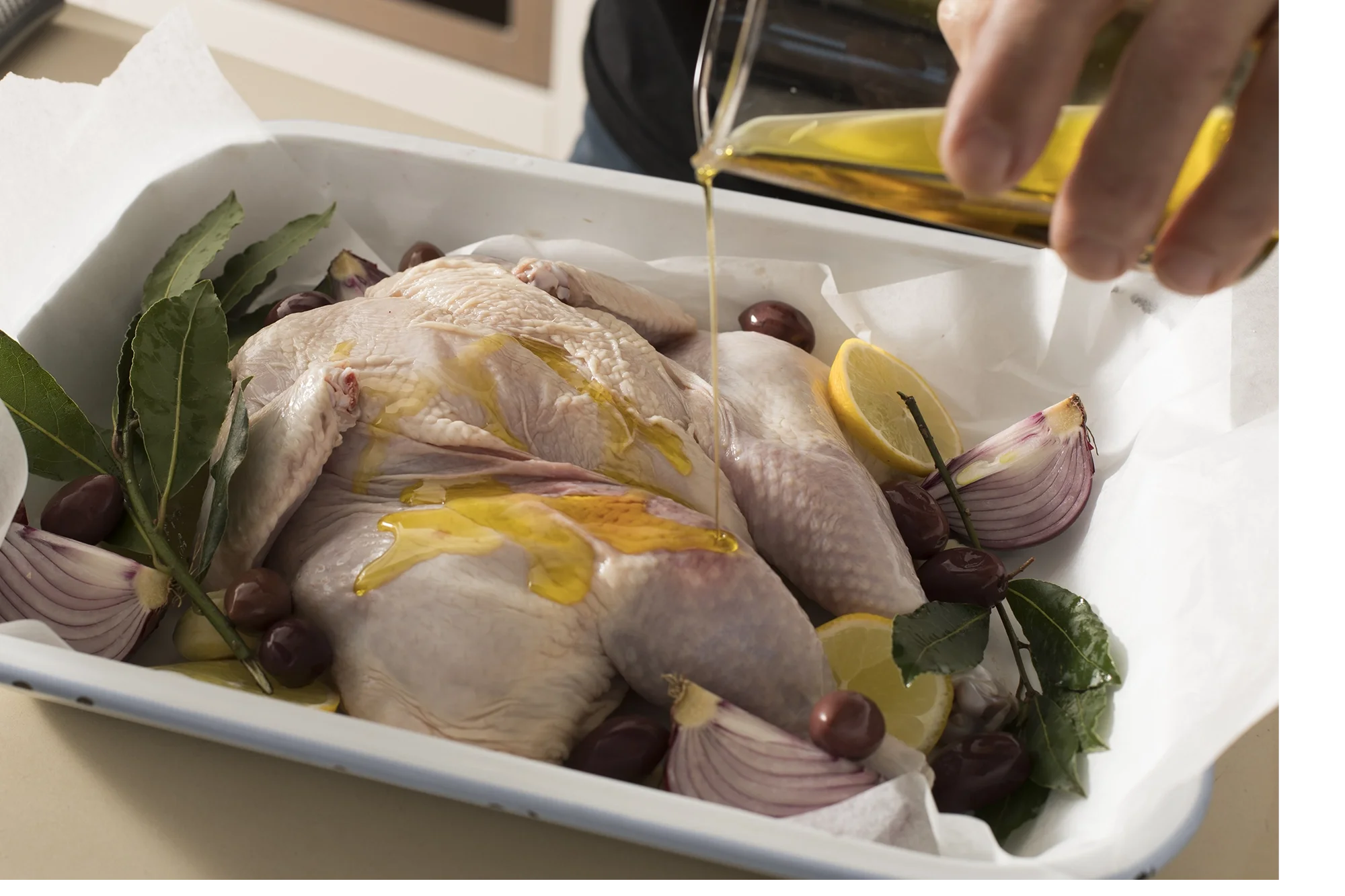 Raw chicken on a baking tray with lemon, garlic, olives, herbs, and olive oil being poured over it.