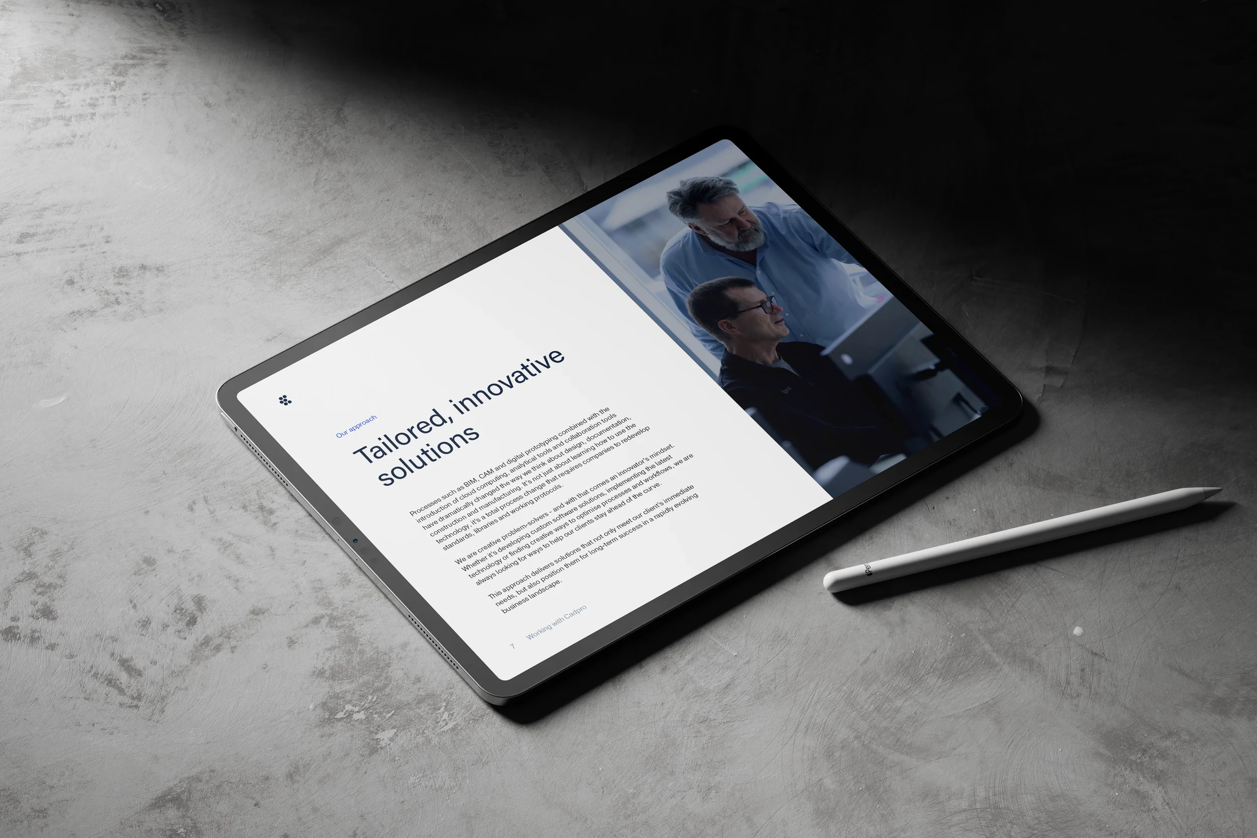 A tablet displaying a webpage titled "Tailored, innovative solutions" with a photo of two men working together, one with a beard and glasses and another with gray hair, in a professional setting. A stylus pen is beside the tablet on a concrete surfac