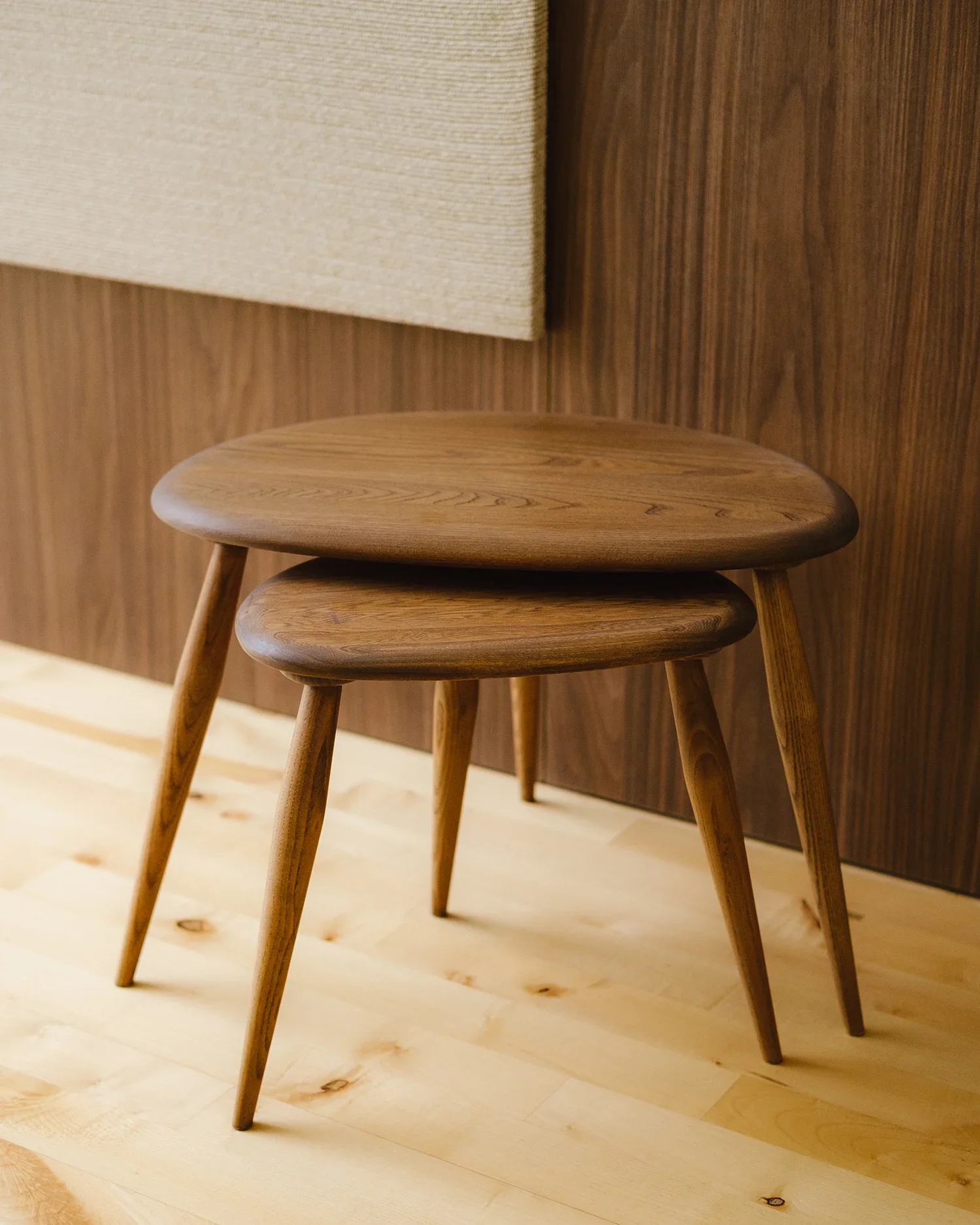 Three wooden stools with rounded seats and angled legs are placed in front of a wood-paneled wall, with a beige fabric-covered object partially visible at the top of the image.
