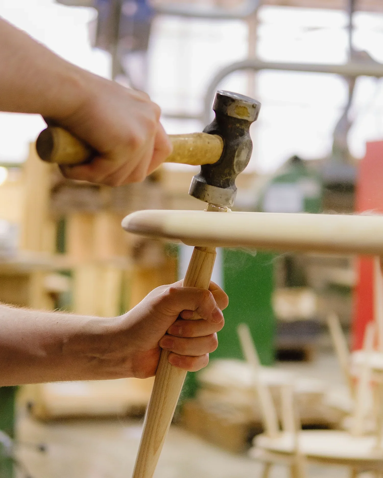 A person holding a rubber mallet and pounding a wooden chair leg in a woodworking workshop with other chairs in the background.
