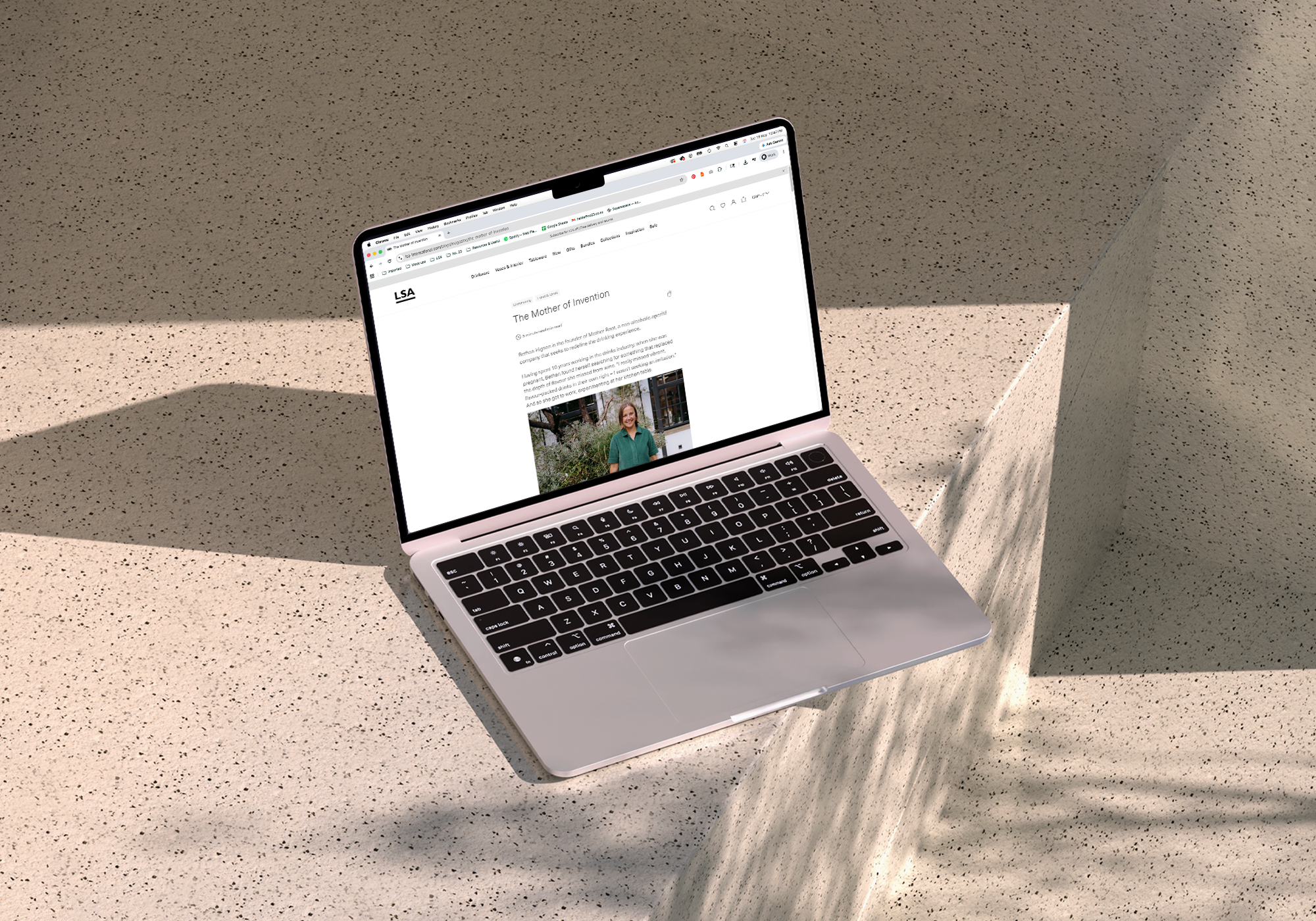 Open laptop on a beige speckled terrazzo surface displaying a web page titled 'The Mother of Invention' with a woman photo and text.