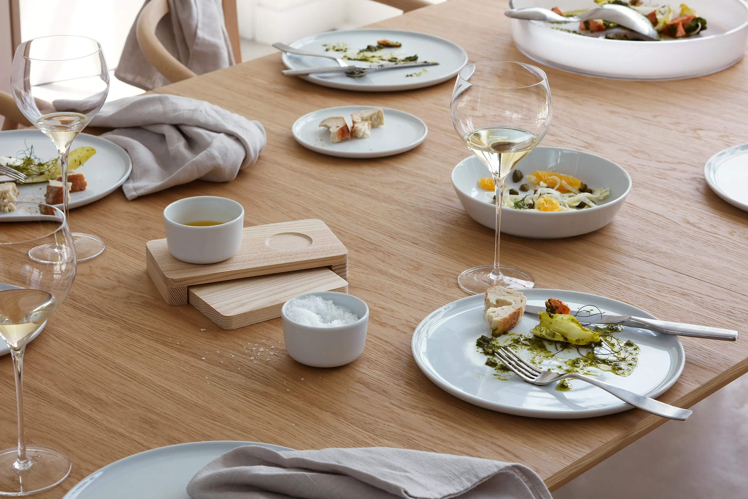 Partially eaten plates of salad and bread, empty wine glasses, a small bowl of salt, a small bird of oil, and a wooden serving board on a wooden dining table after a meal.