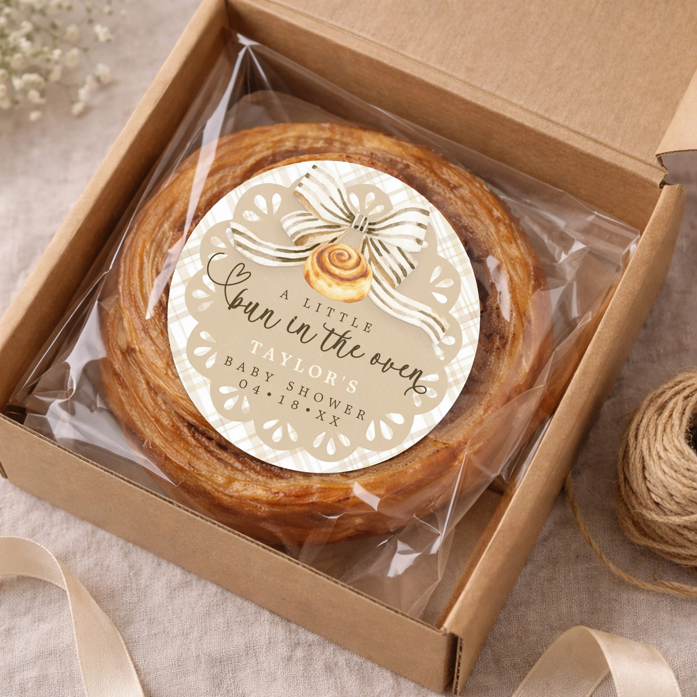 Bun in the Oven baby shower favor sticker on cinnamon roll inside kraft bakery box with clear wrap, styled with ribbon and neutral decor.