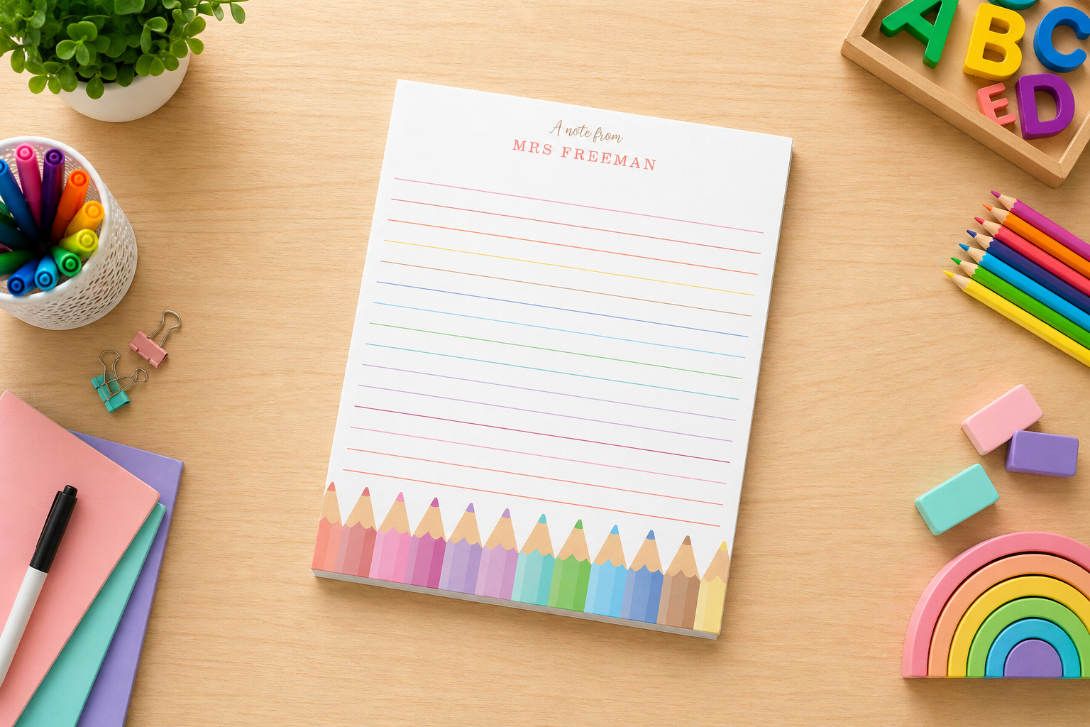 Personalized lined teacher notepad on a wooden desk featuring rainbow pencil artwork, surrounded by markers, pencils, pastel erasers, notebooks, alphabet blocks, and a rainbow toy.