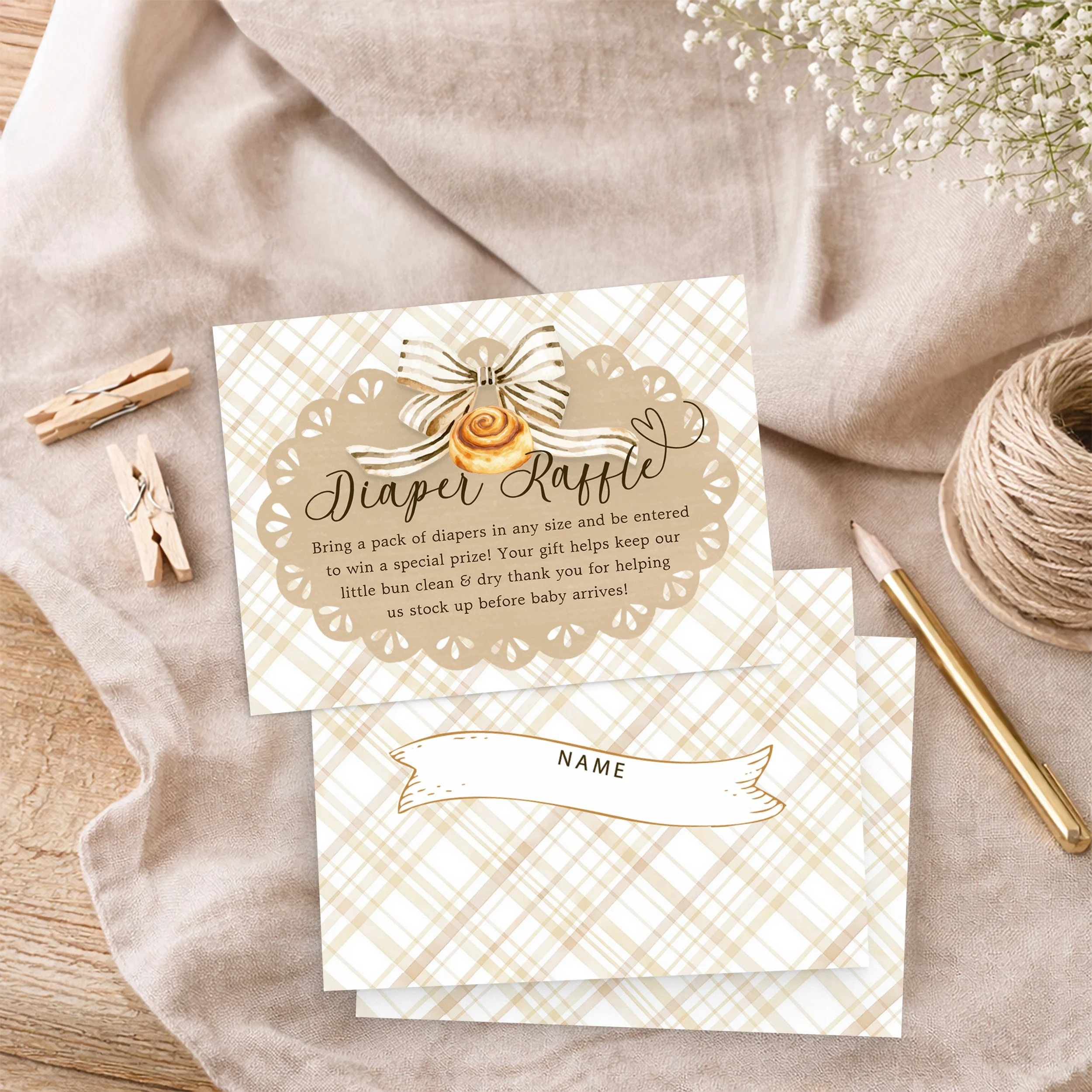 Bun in the Oven baby shower diaper raffle cards with neutral gingham design, displayed on soft fabric with twine, pencil, and wooden clothespins.