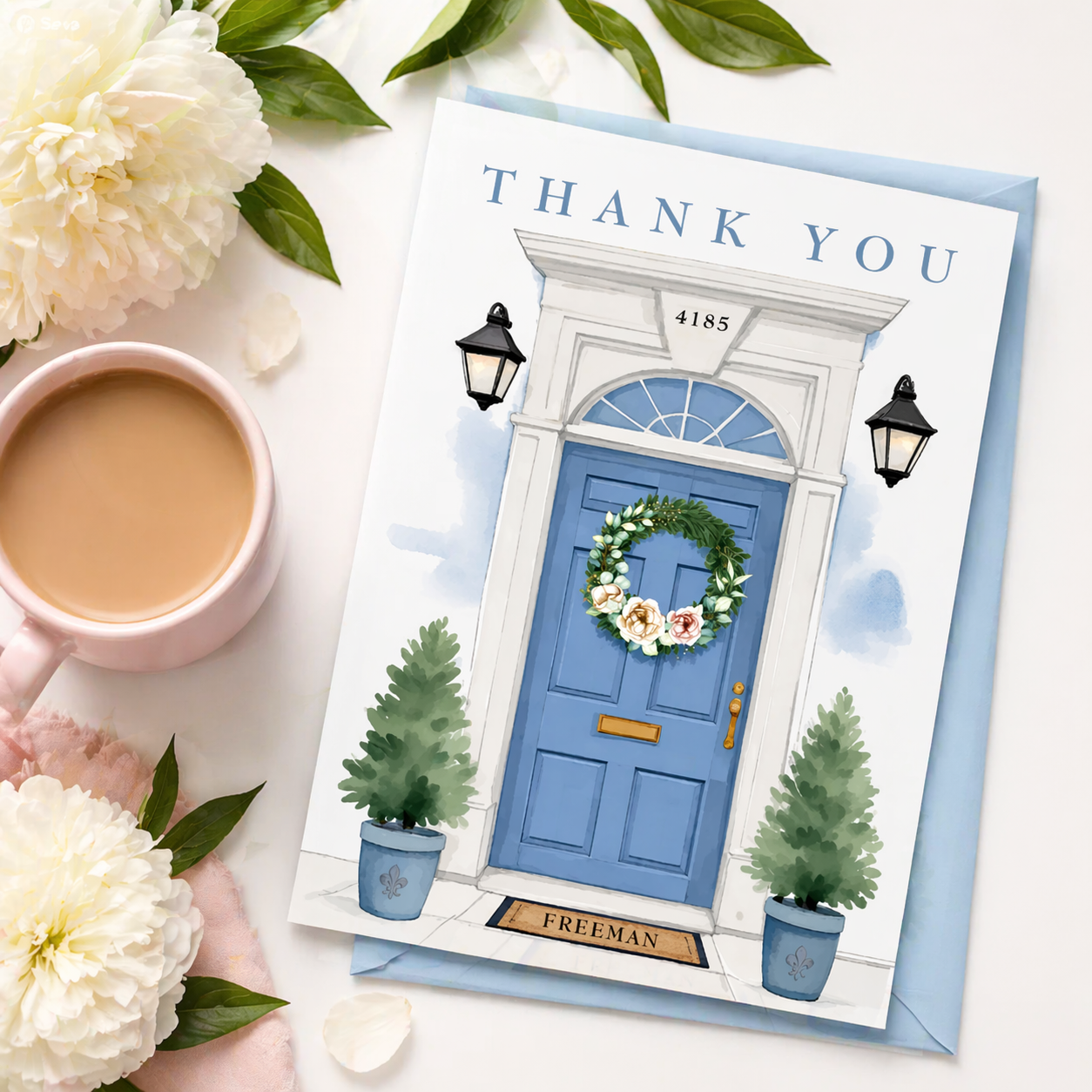 Flat lay styled image of a personalized housewarming thank you card featuring a watercolor blue front door with floral wreath, paired with a soft blue envelope, white peonies, greenery, and a pink coffee cup on a bright neutral background.