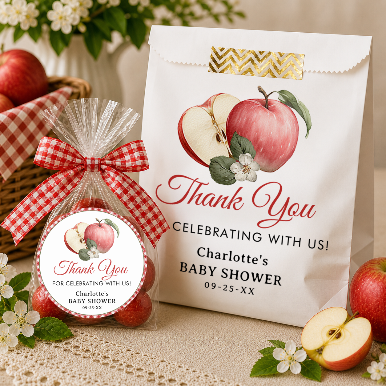 Apple-themed baby shower favors displayed on a neutral tabletop, including a personalized white thank-you favor bag and a clear cello bag filled with red apples tied with a red gingham bow.