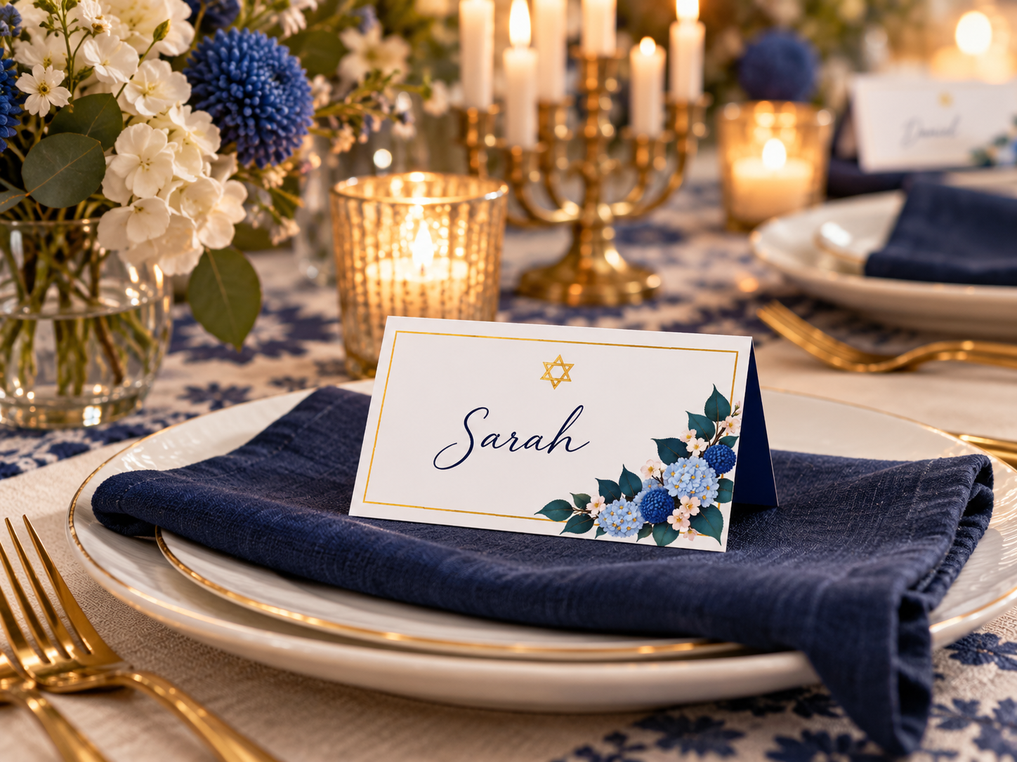 Elegant Hanukkah dinner place setting featuring a personalized floral place card with handwritten guest name, gold Star of David, navy linen napkin, gold-rimmed plates, and blue and white flowers.