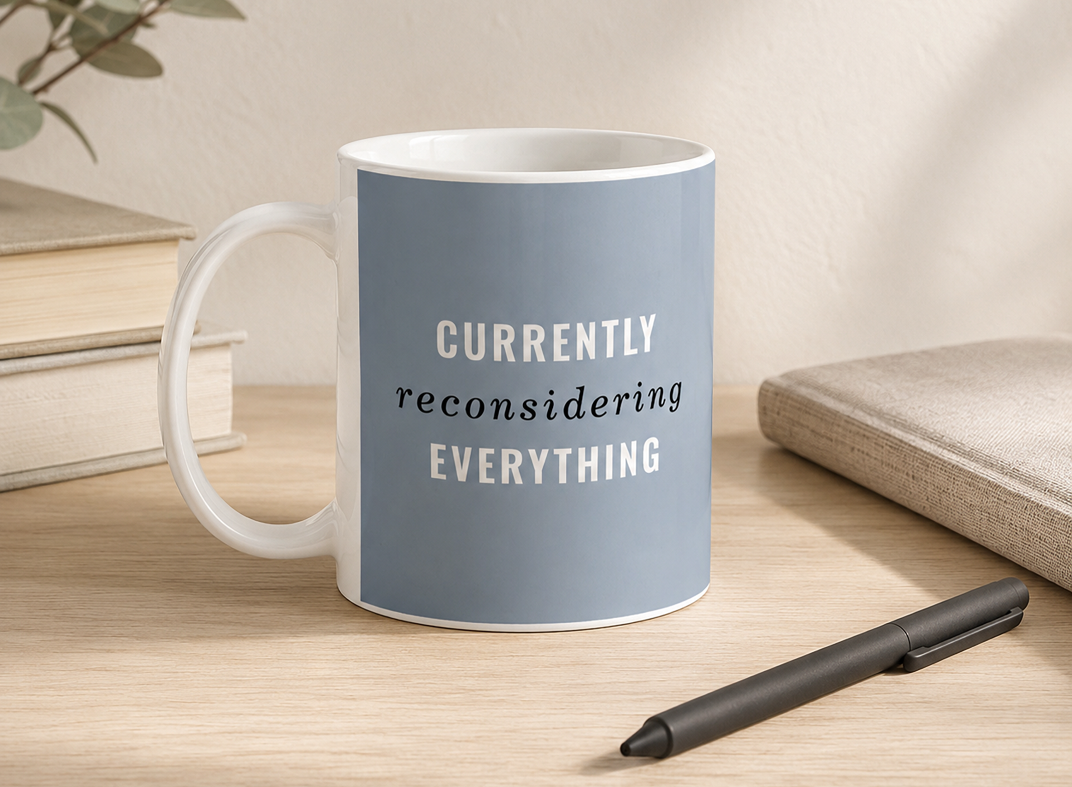 Dusty blue coffee mug reading “Currently Reconsidering Everything” on a pale wood desk with a notebook and pen.