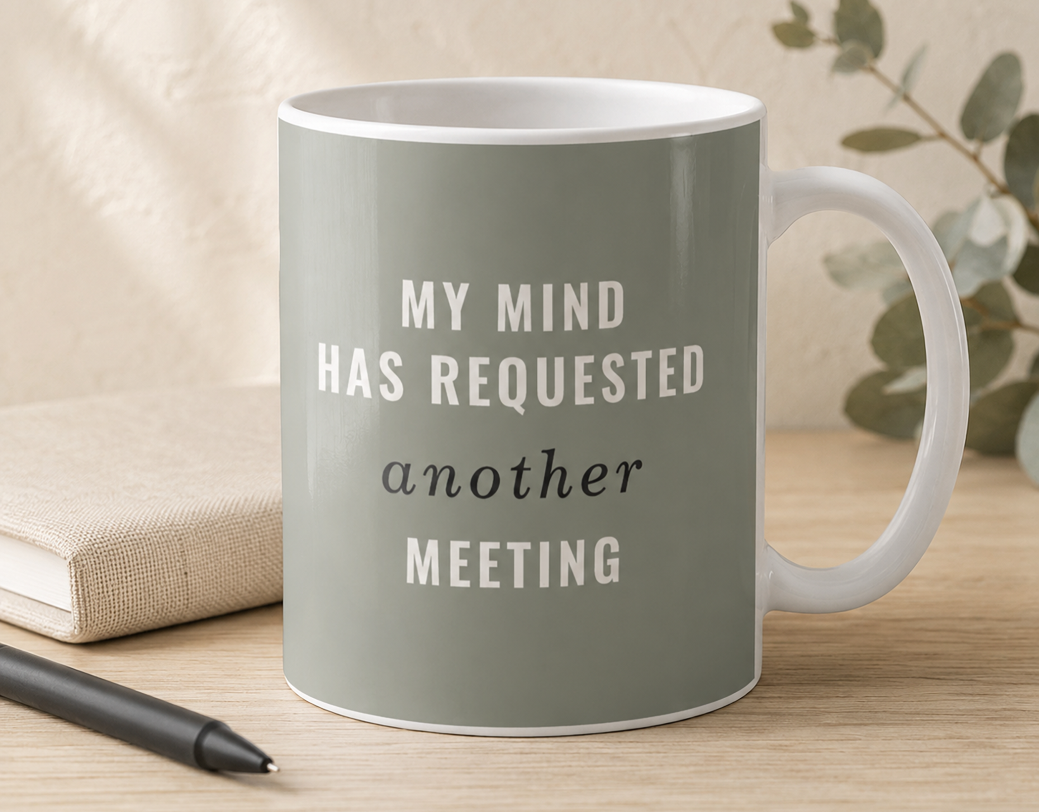 Sage green coffee mug reading “My Mind Has Requested Another Meeting” styled with a notebook and pen on a warm neutral desk.