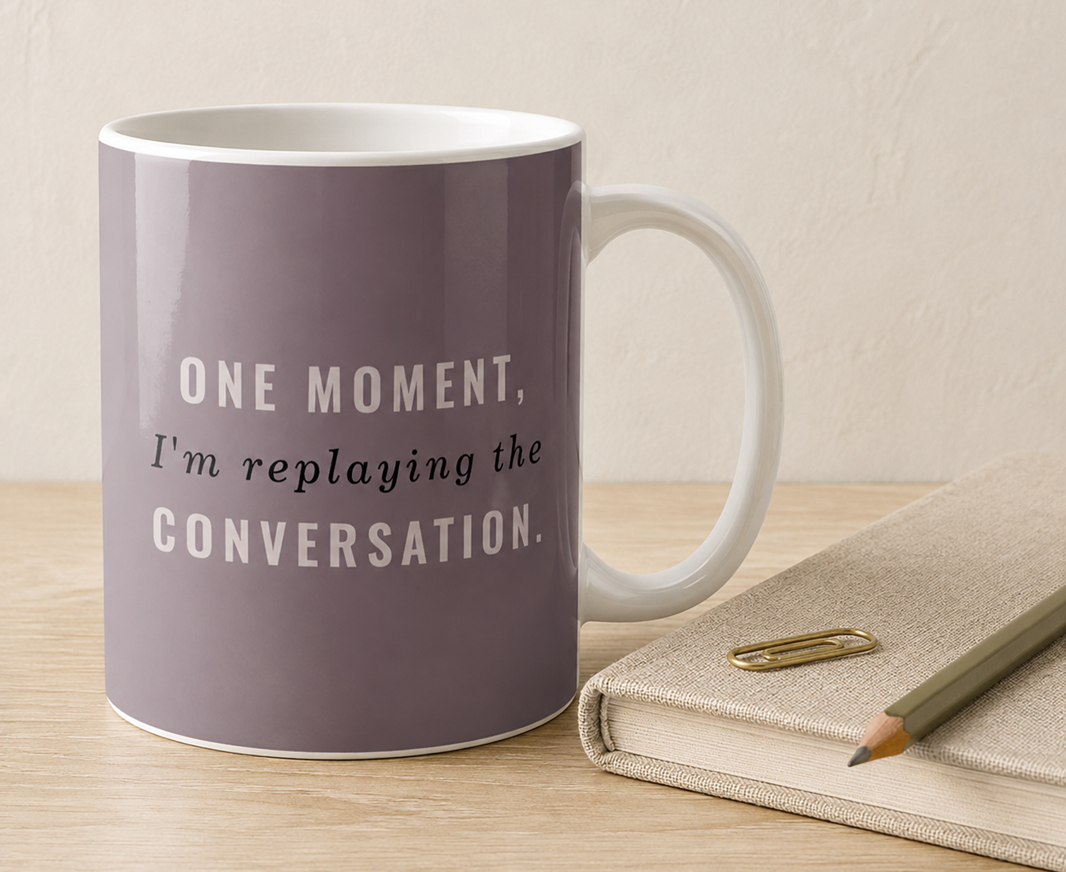 Muted mauve coffee mug reading “One Moment, I’m Replaying the Conversation.” beside a notebook and pencil on a warm desk.