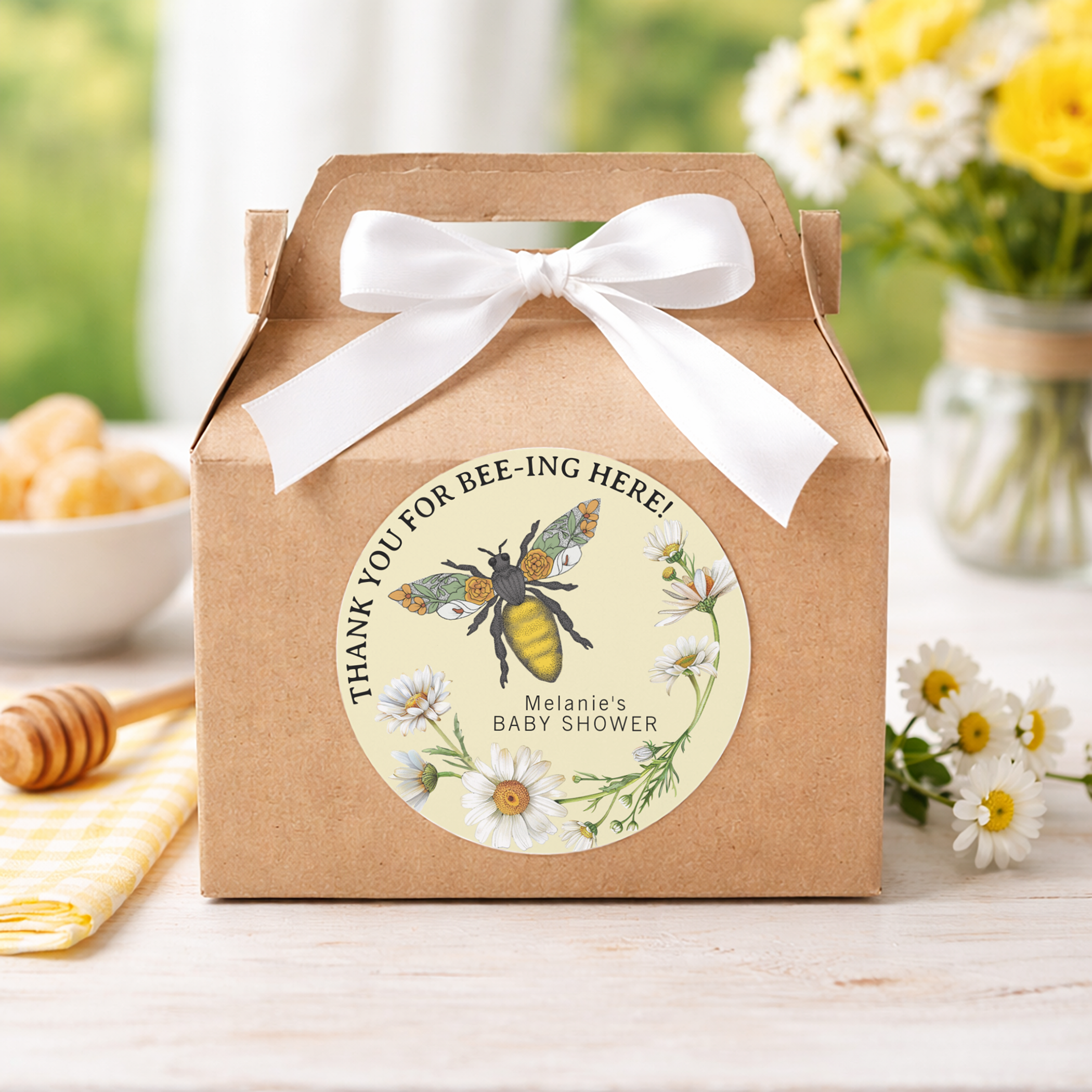Kraft paper baby shower favor box with a white ribbon bow and honey bee themed “Thank You for Bee-ing Here” sticker, styled with daisies, honey dipper, and soft yellow spring décor.