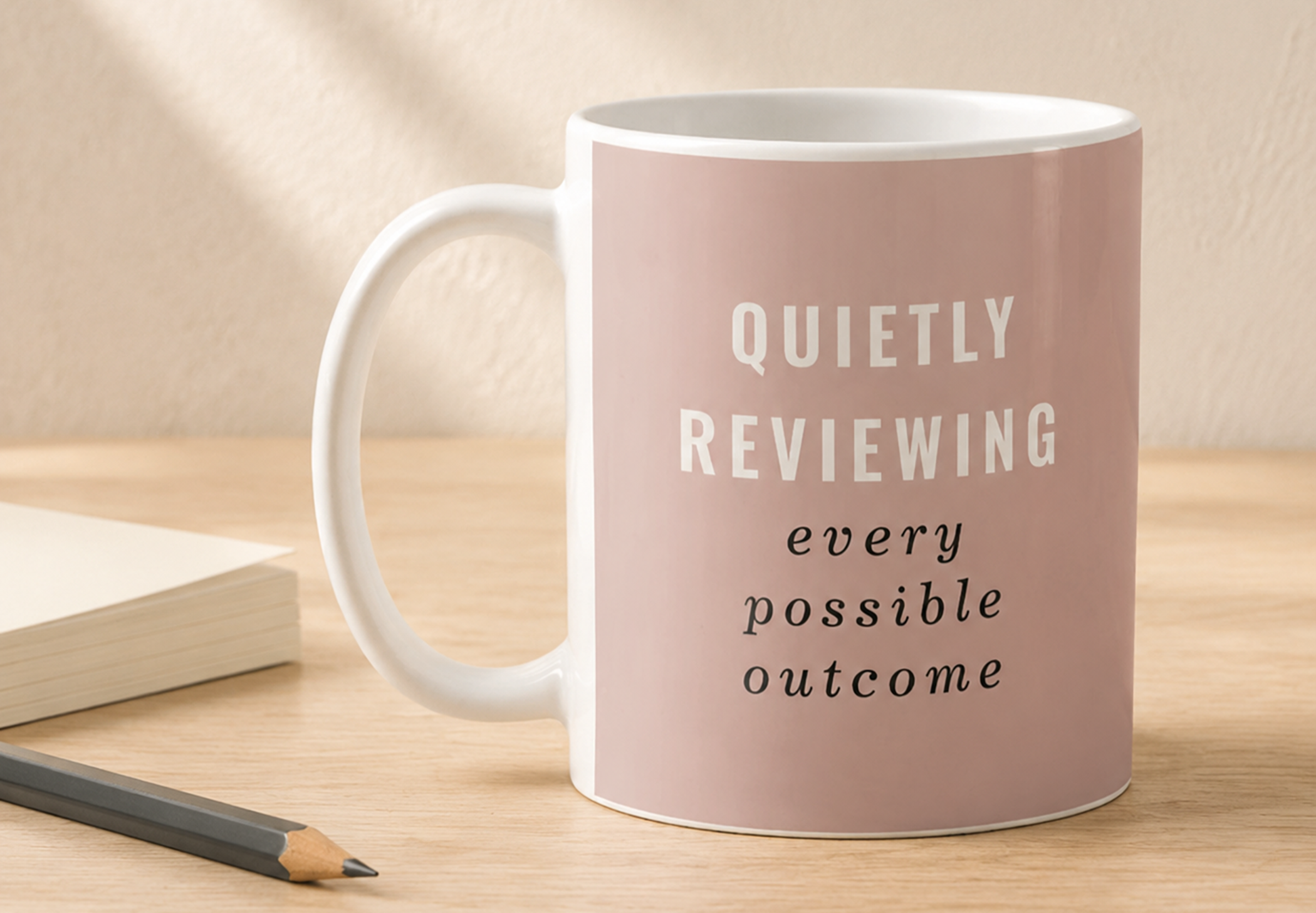 Blush pink coffee mug reading “Quietly Reviewing Every Possible Outcome” styled with an open notebook and pencil on a pale wood desk.