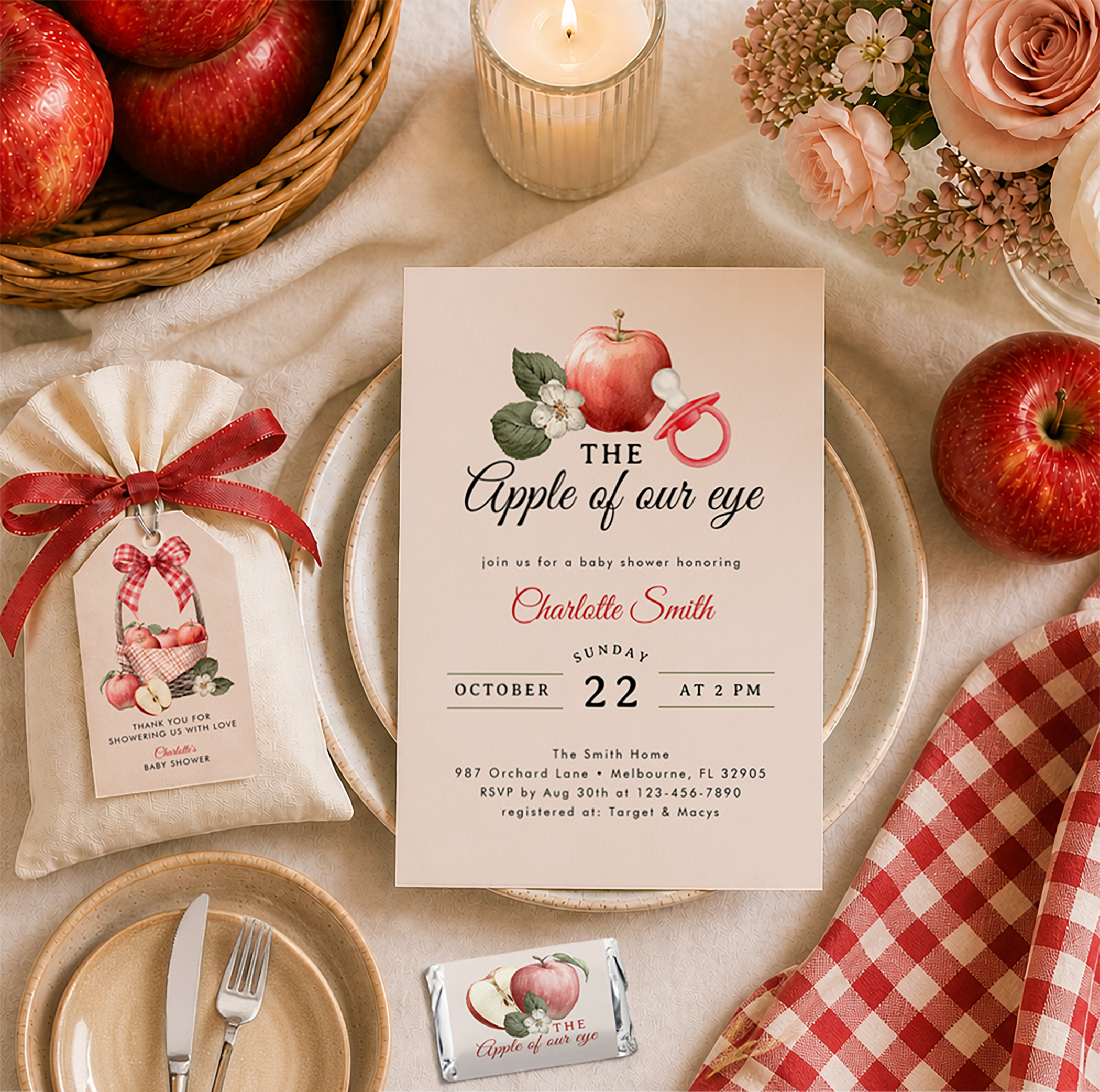 Flat lay apple-themed baby shower tablescape featuring a white invitation card, cream favor bag with gift tag, wrapped candy favor, red gingham napkin, fresh red apples, blush and cream roses, and a lit candle in warm neutral tones.