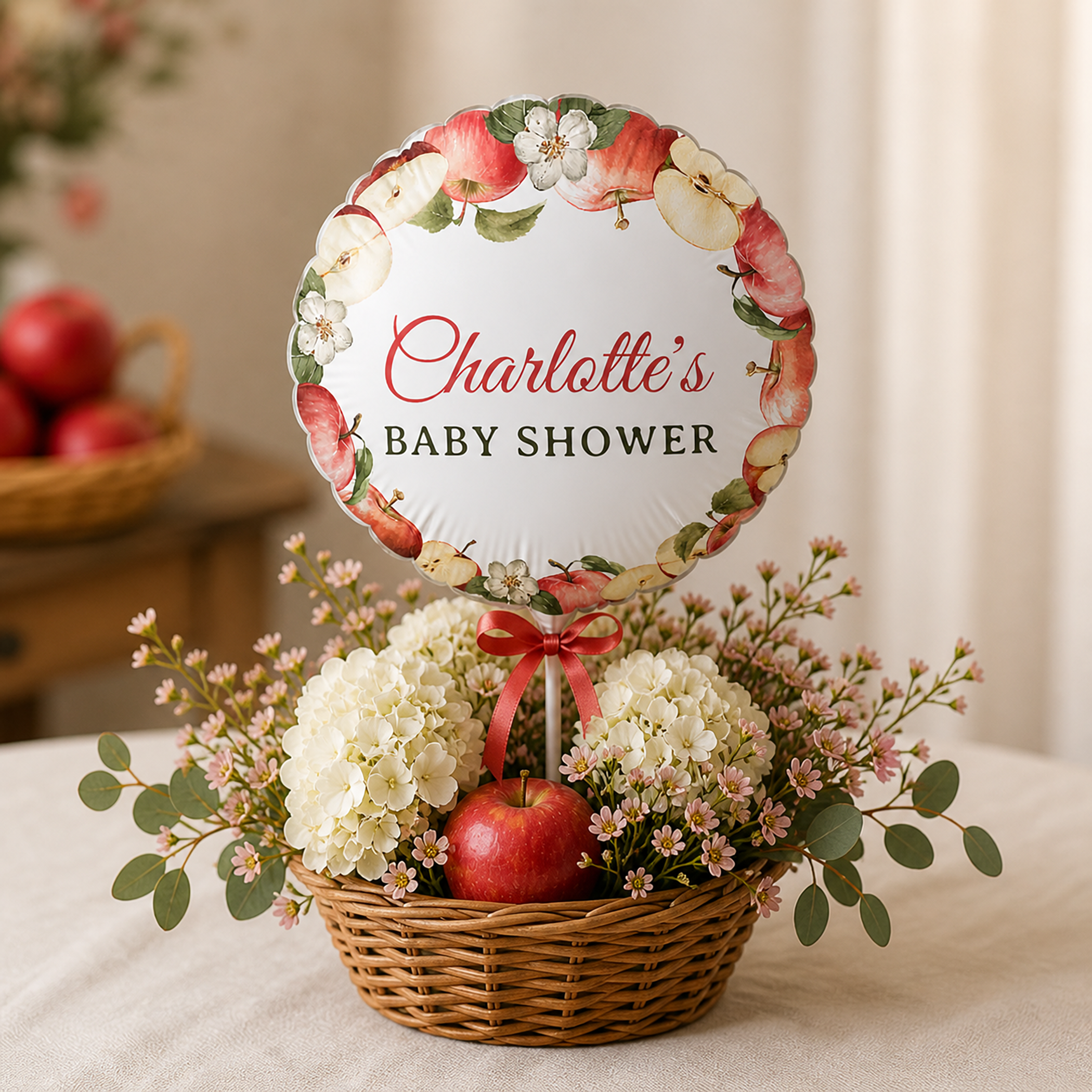 Round personalized baby shower balloon with apple blossom design displayed in a woven basket centerpiece filled with white flowers, pink blooms, greenery, and a red apple on a neutral table setting.