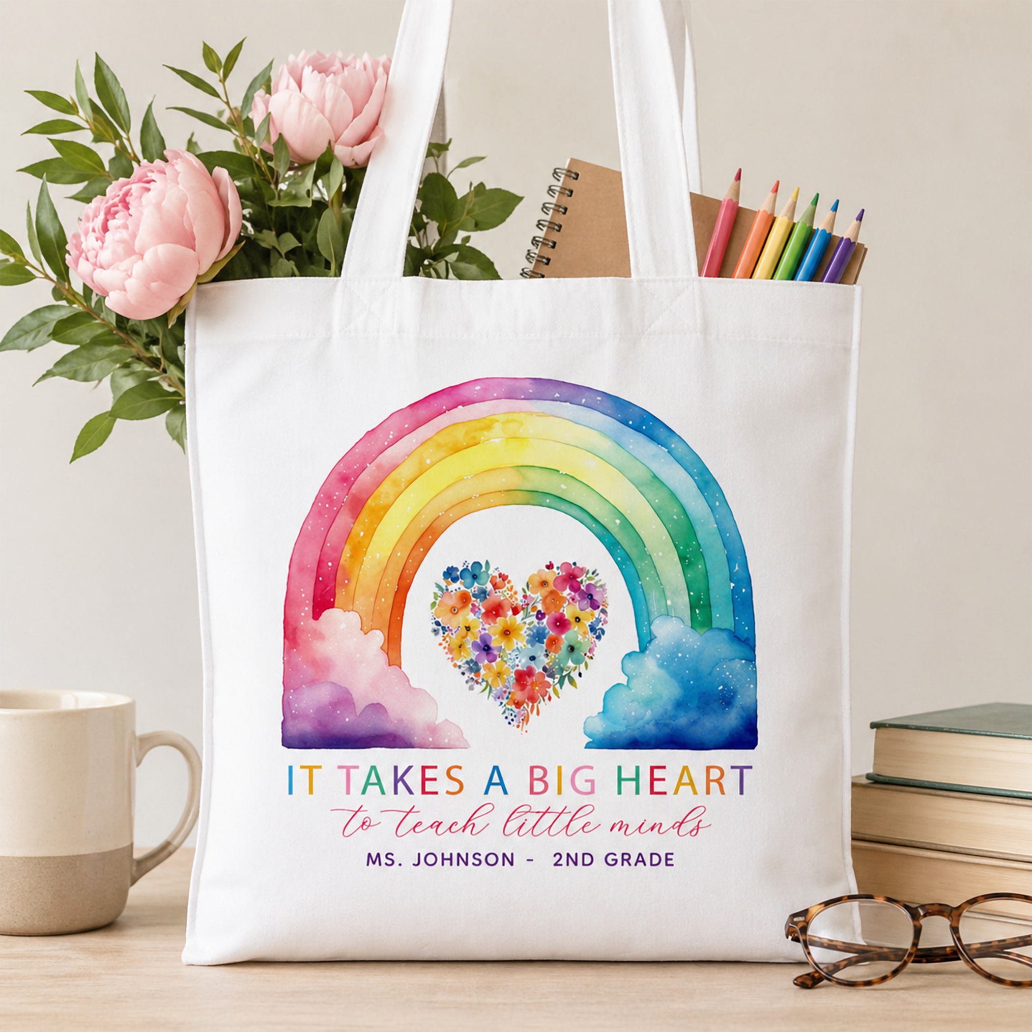 Personalized white teacher tote bag with colorful rainbow and floral heart design, displayed on a desk with flowers, pencils, books, coffee mug, and glasses in a bright classroom-inspired setting.