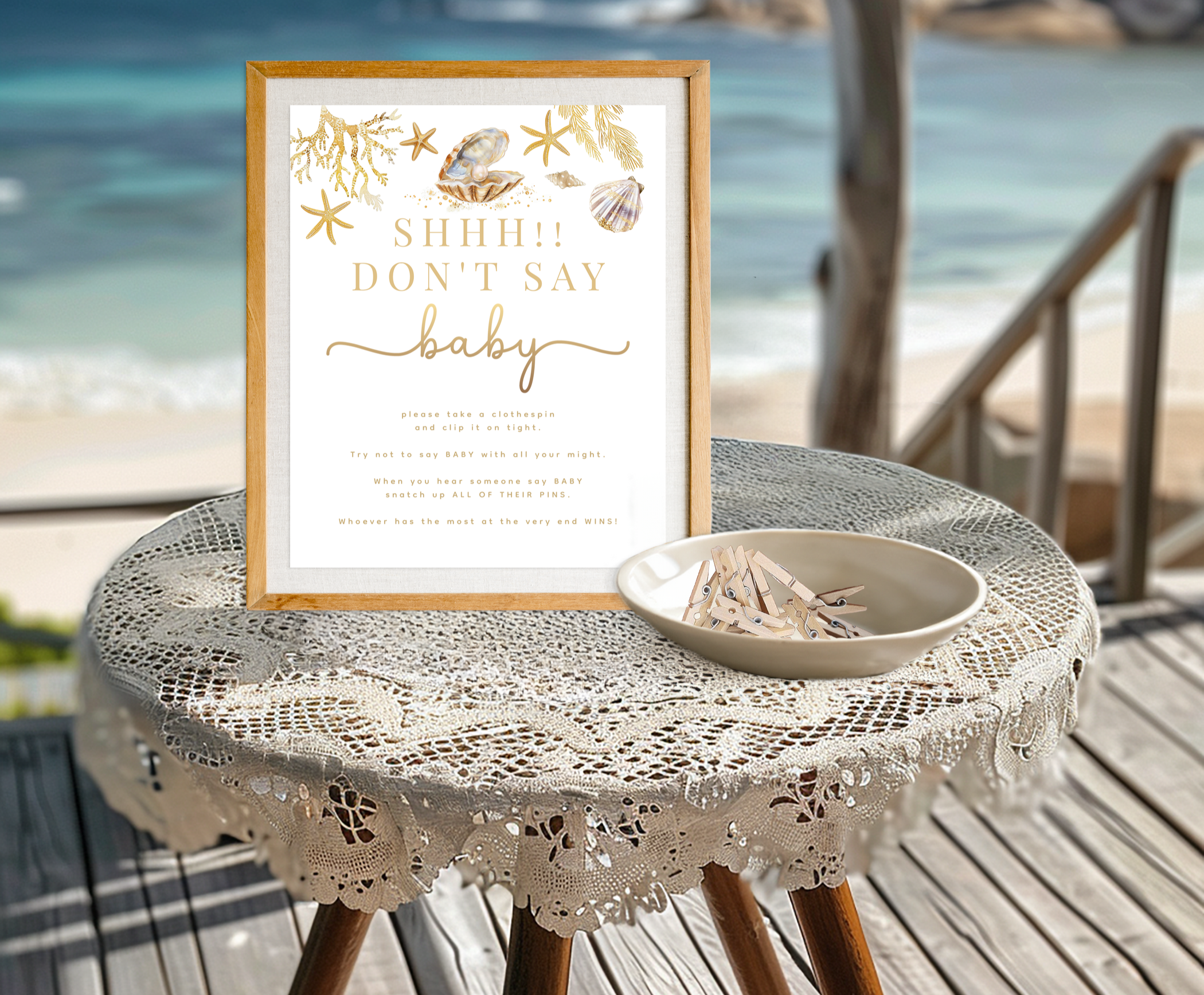 Beach-themed Don't Say Baby baby shower game sign displayed in a wooden frame with clothespins on a lace-covered table overlooking the ocean.