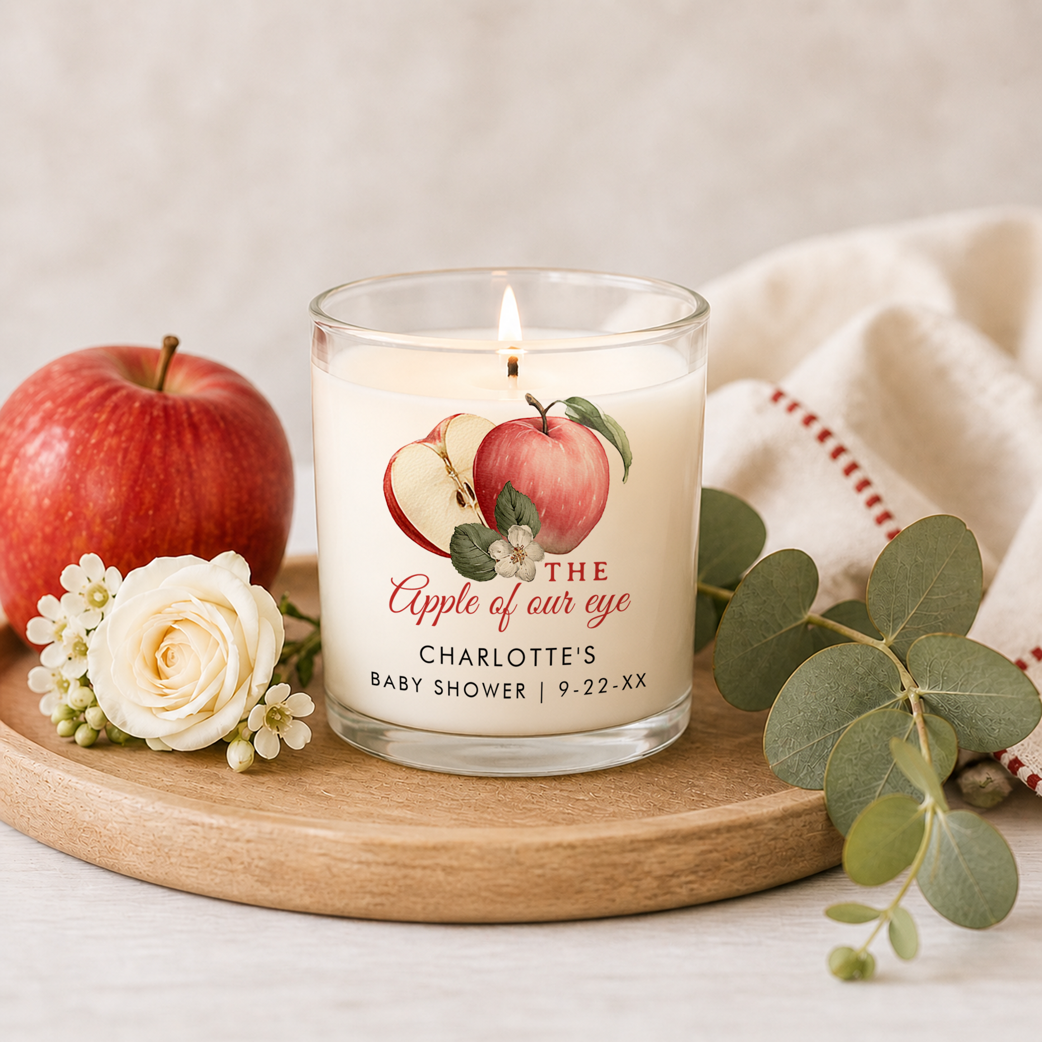 Personalized baby shower candle in a clear glass jar reading “The Apple of Our Eye Charlotte’s Baby Shower” displayed on a wooden tray with a red apple, white rose, small blossoms, eucalyptus stems, and soft neutral fabric background.