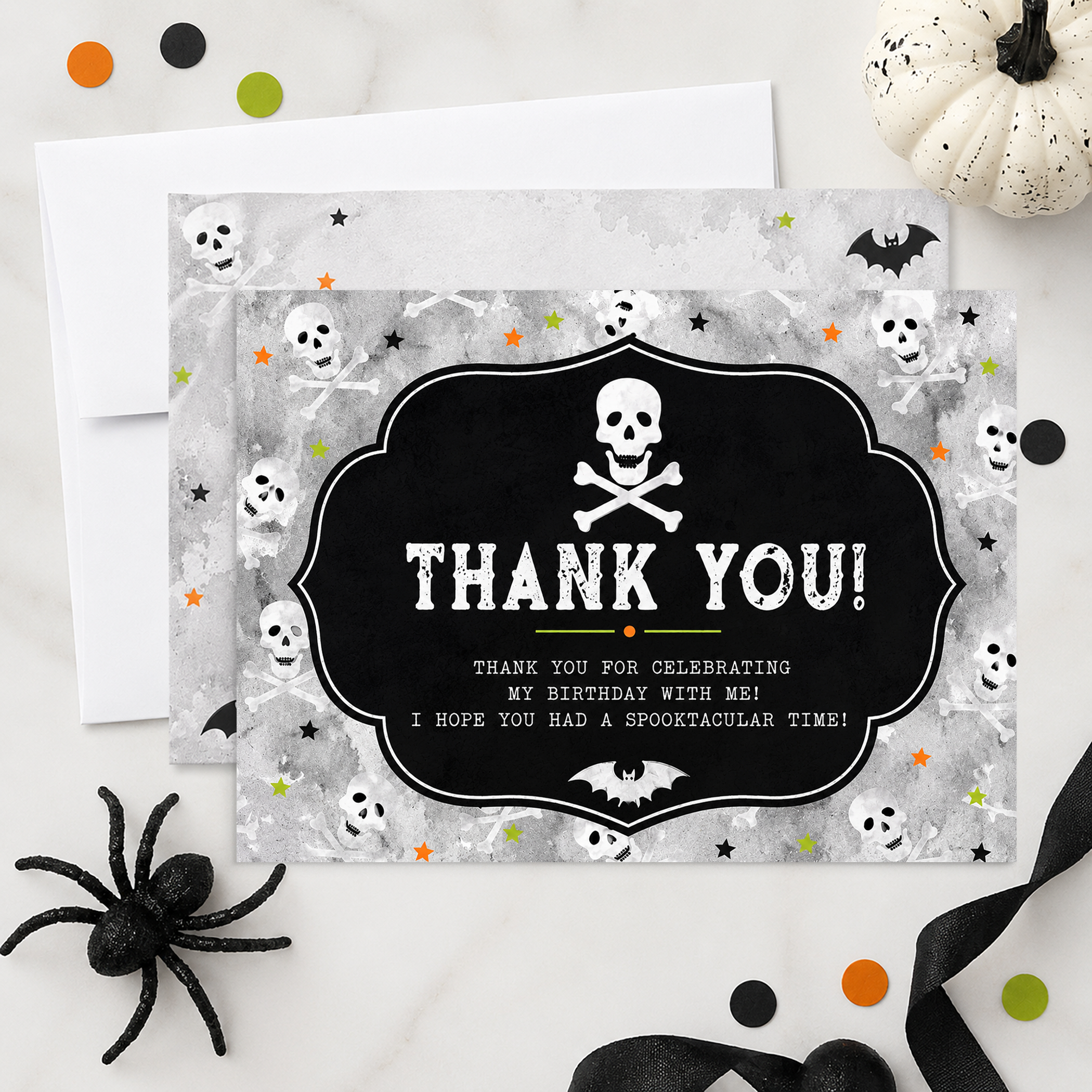 Halloween birthday thank you flat card with skull and crossbones design styled with envelope, black ribbon, spider decor, and festive orange and green confetti on a marble background.