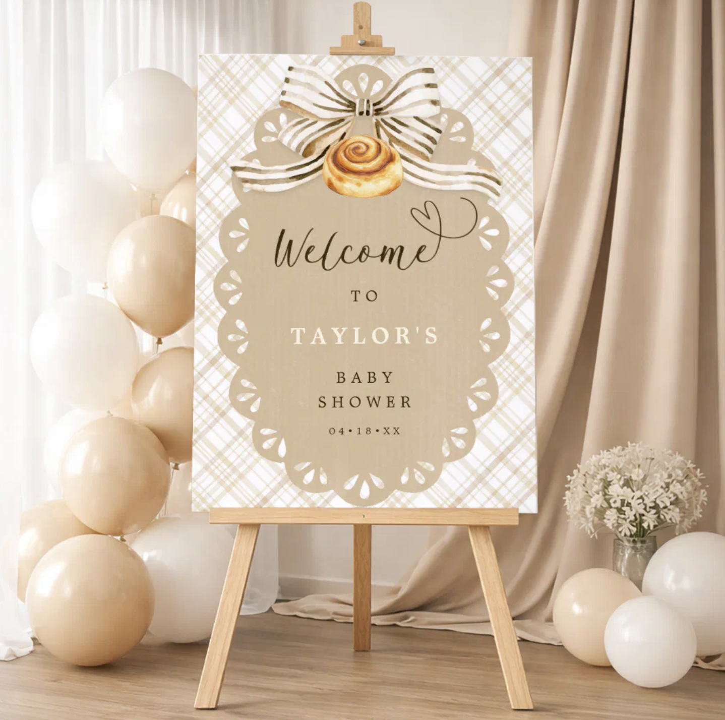 Neutral Bun in the Oven baby shower welcome sign on wooden easel with beige gingham background, cinnamon roll illustration, striped bow, cream balloons, draped fabric backdrop, and floral accents.