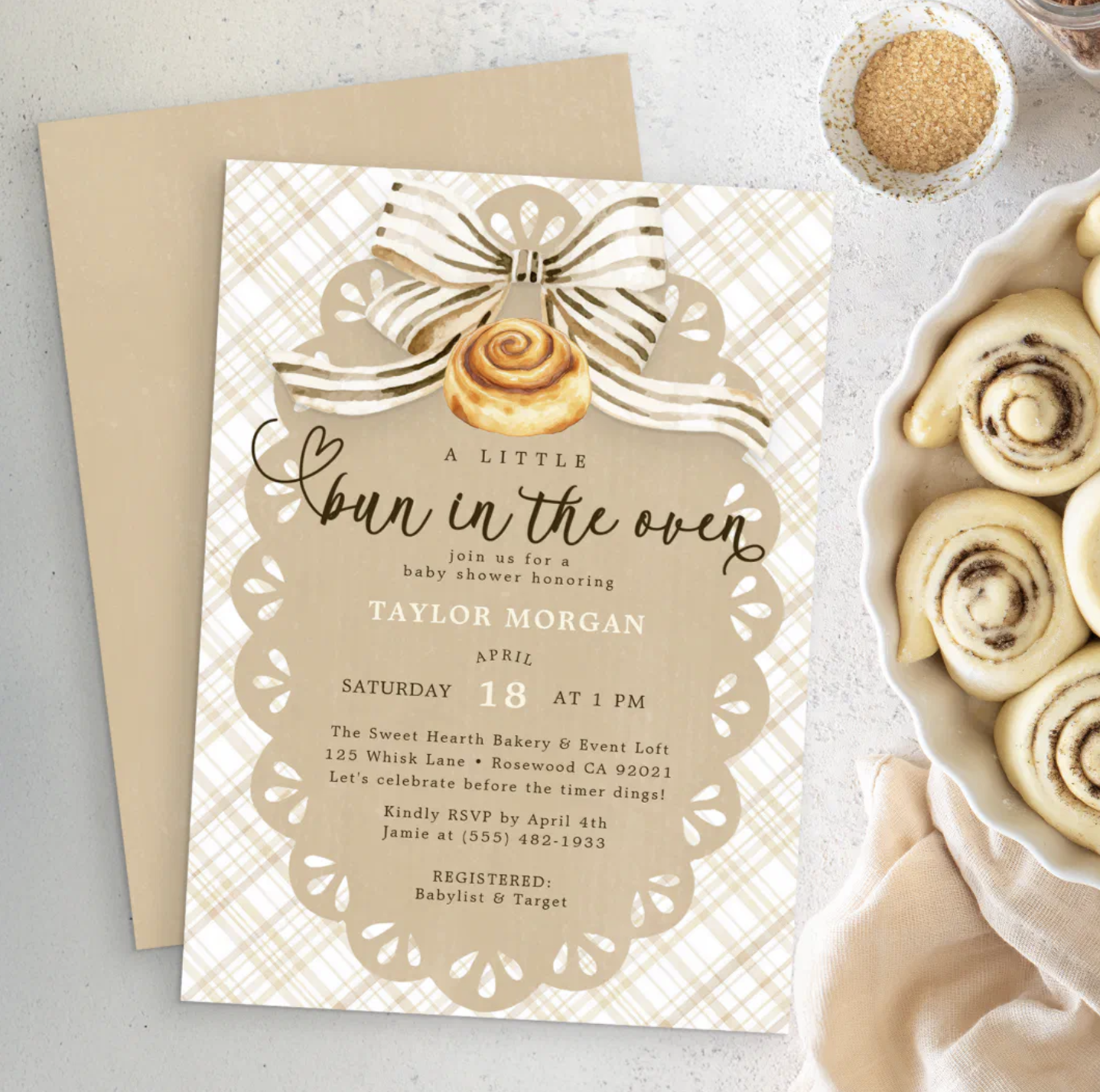 Neutral Bun in the Oven baby shower invitation with beige gingham background, cinnamon roll illustration, striped bow, kraft envelope, and cinnamon rolls beside the invitation.