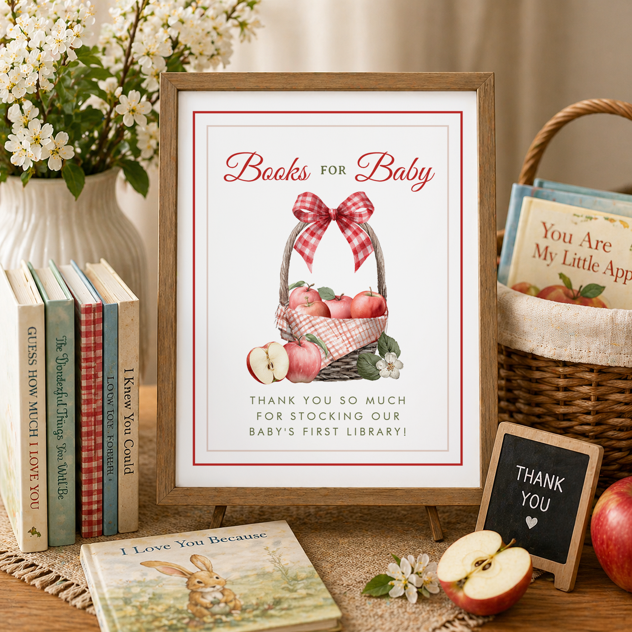 Rustic Books for Baby baby shower table featuring framed apple-themed sign, stacked children’s books, wicker basket of books, blooming white flowers, apples, and small thank-you chalkboard sign in warm neutral setting.