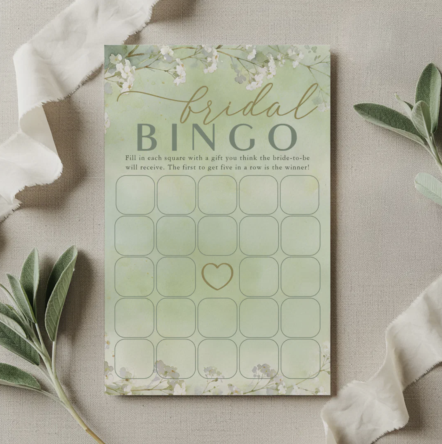 Elegant bridal bingo game card with soft sage green watercolor background, delicate floral accents, and bingo grid displayed on neutral linen fabric with greenery. Printable bridal shower game idea for gift opening entertainment.