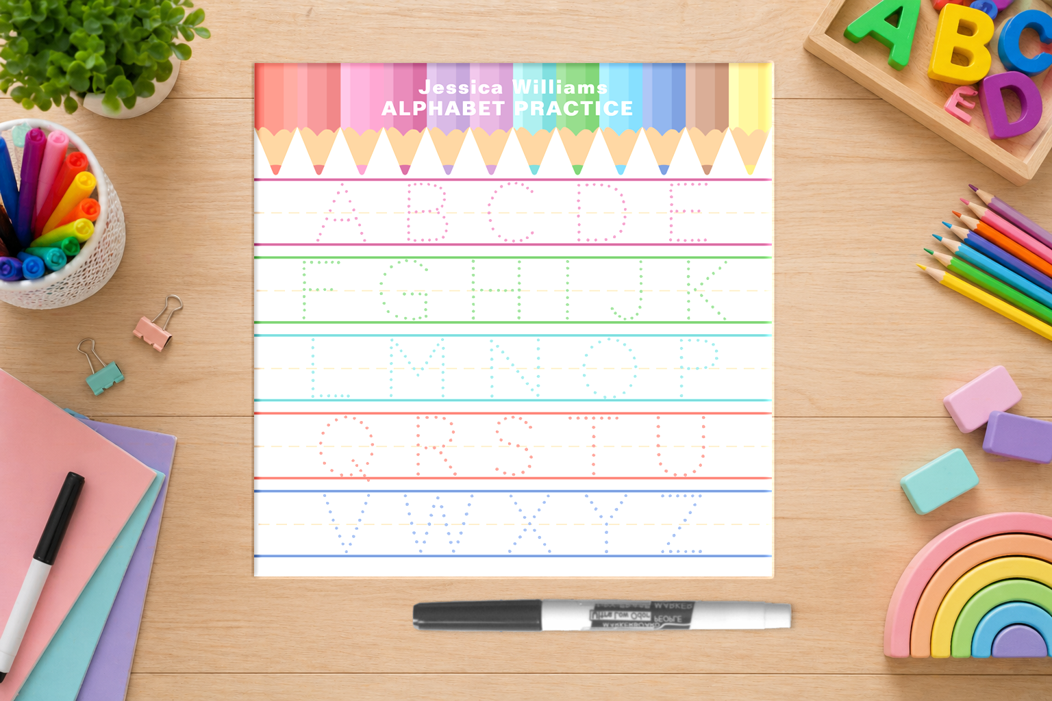 Personalized square alphabet tracing dry erase board on a wooden desk surrounded by markers, pencils, erasers, and rainbow toys, featuring dotted uppercase letters A through Z for handwriting practice.