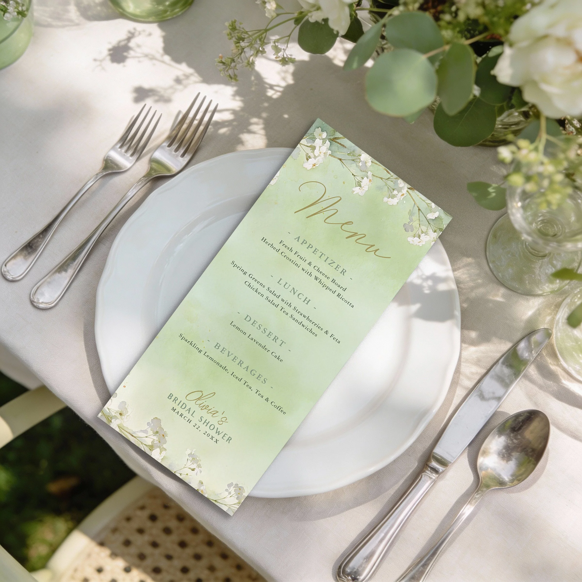 Sage green floral bridal shower menu card on an elegant table setting with white plates and garden sunlight.