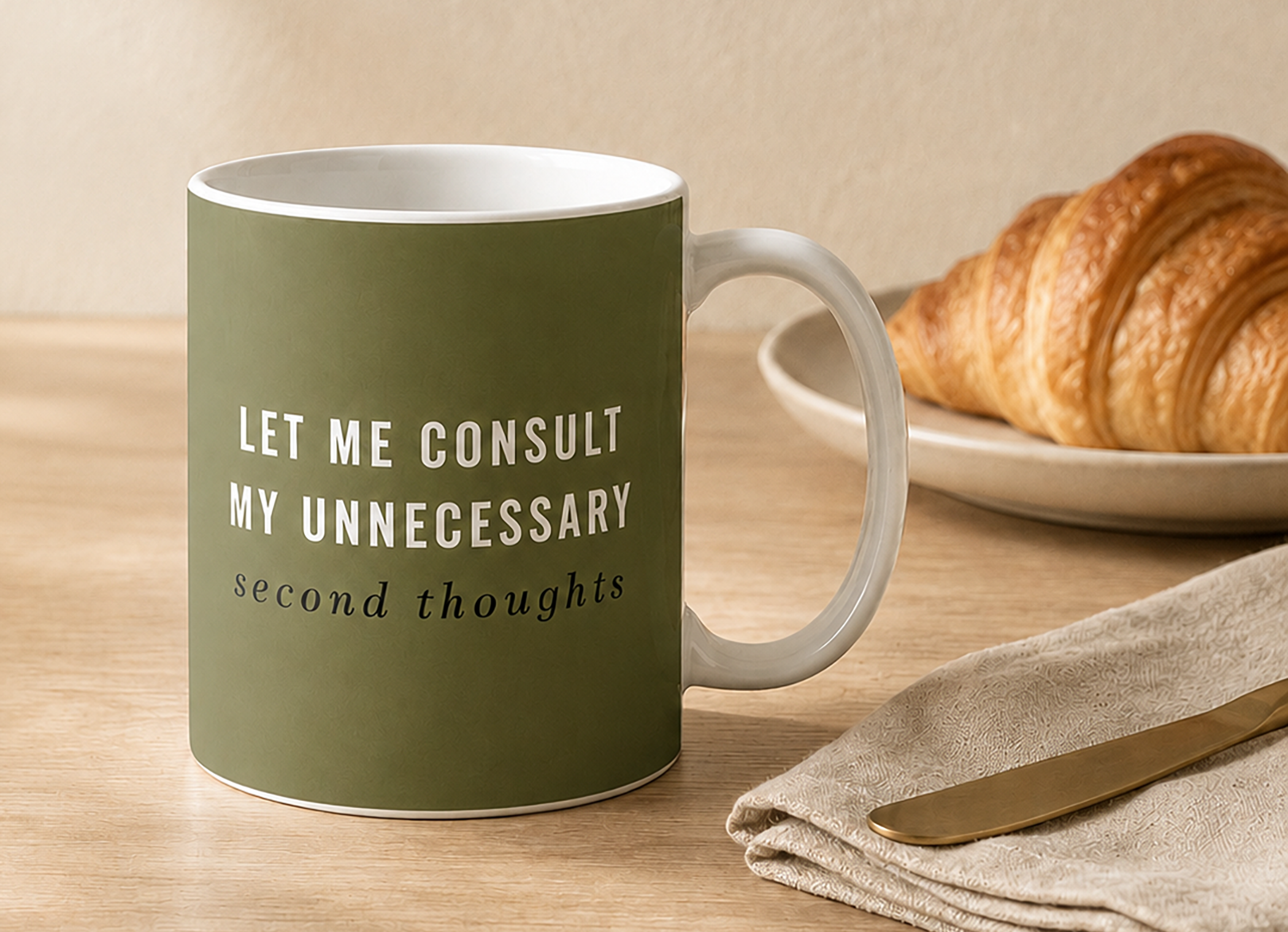 Olive green coffee mug reading “Let Me Consult My Unnecessary Second Thoughts” on a breakfast-inspired neutral tabletop with a croissant.