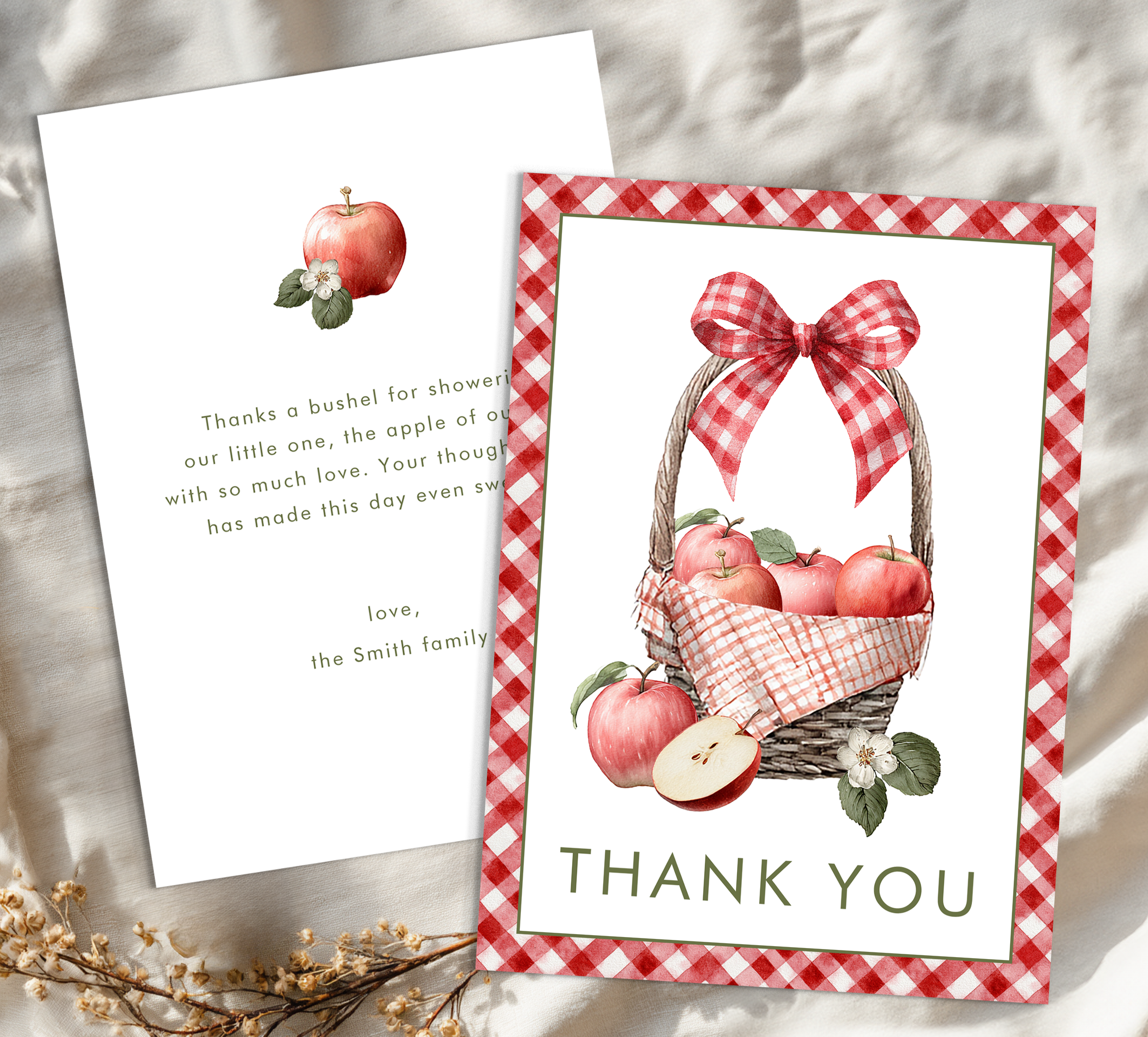 Flat lay of personalized apple themed baby shower thank you cards featuring red gingham border, basket of apples artwork, and matching inside message on neutral fabric background.