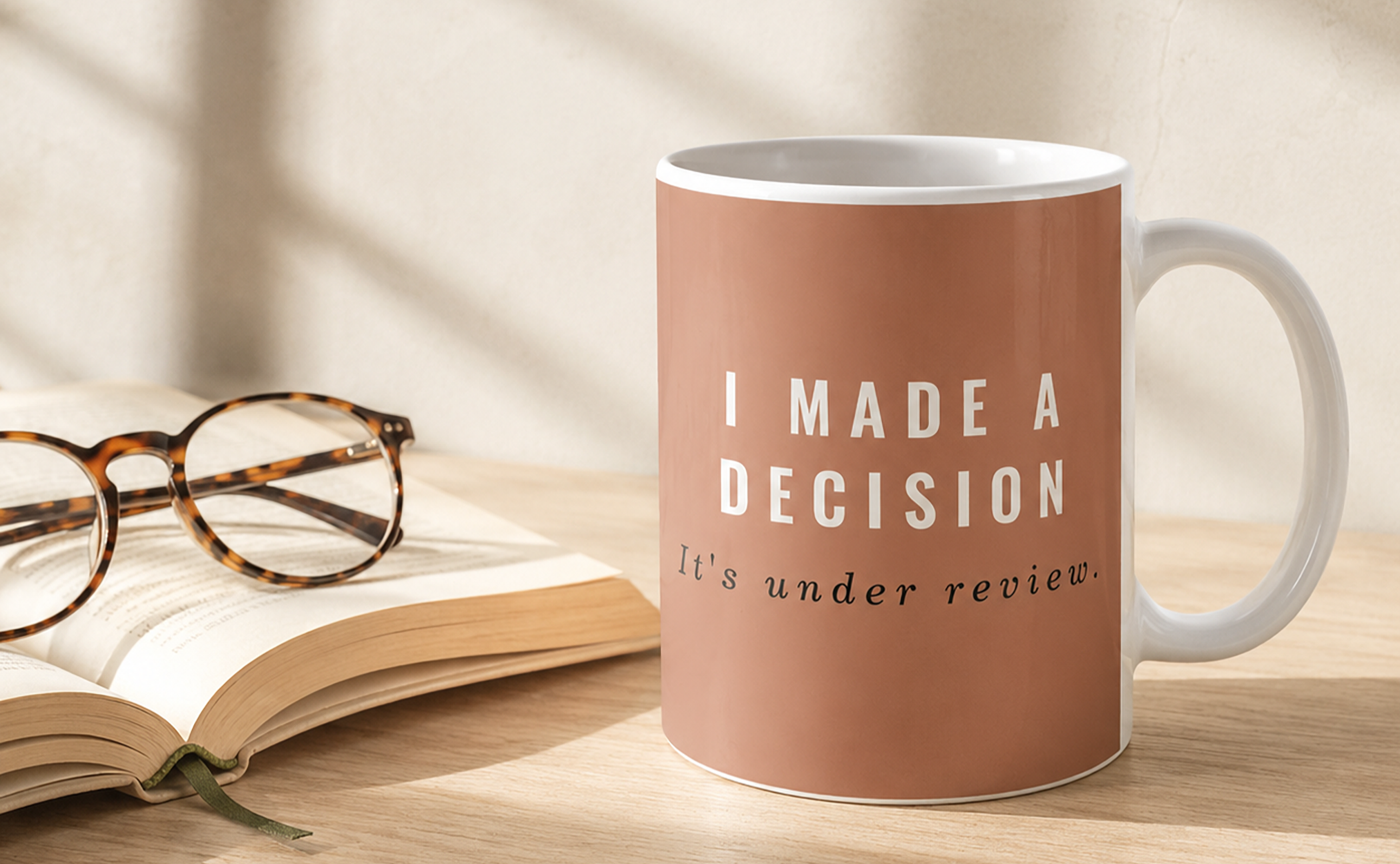 Terracotta coffee mug reading “I Made a Decision. It’s Under Review.” on a softly styled desk with an open book and glasses.