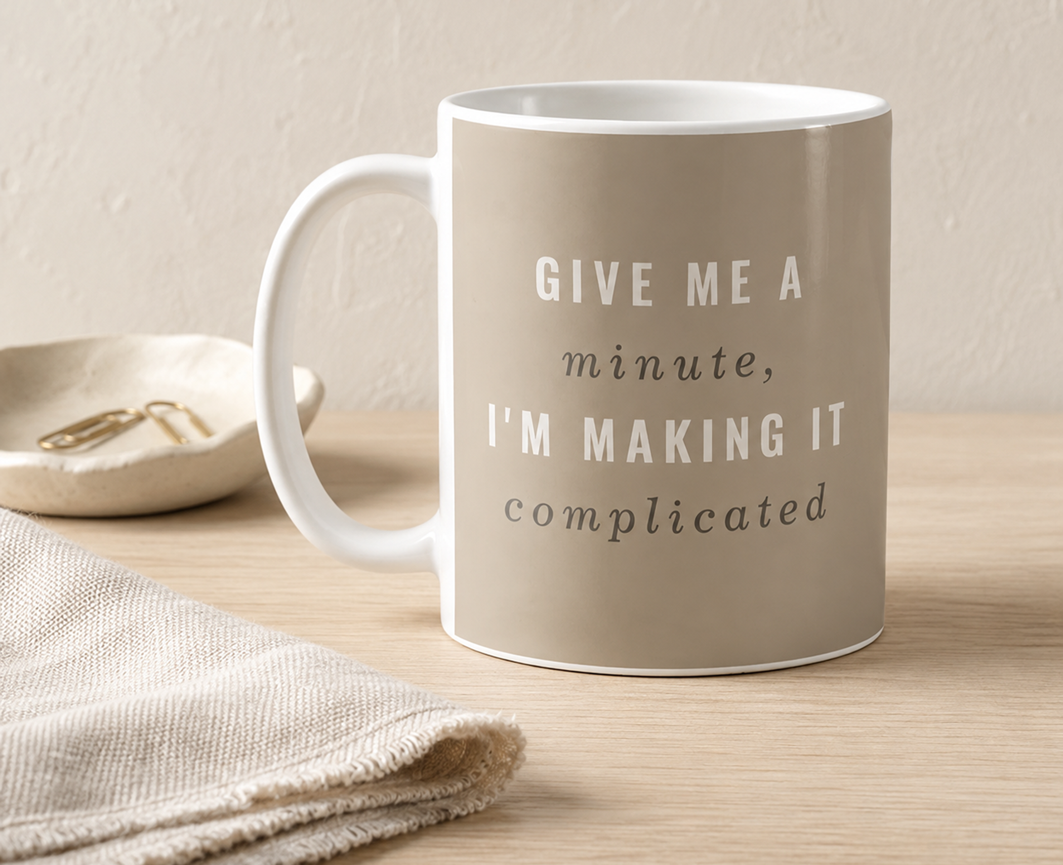 Taupe coffee mug reading “Give Me a Minute, I’m Making It Complicated” styled with neutral linens on a pale wood desk.