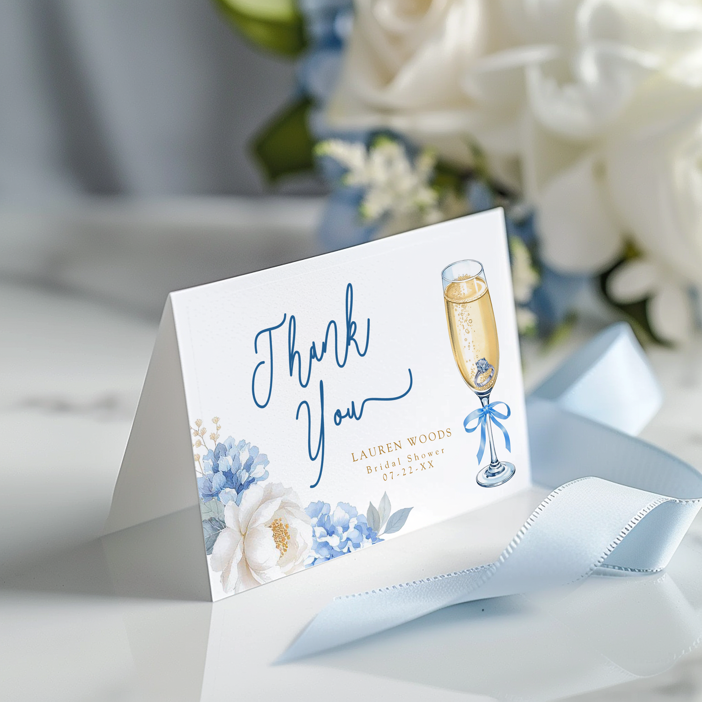 Folded thank you card featuring a champagne flute illustration, soft blue floral accents, and elegant script lettering.