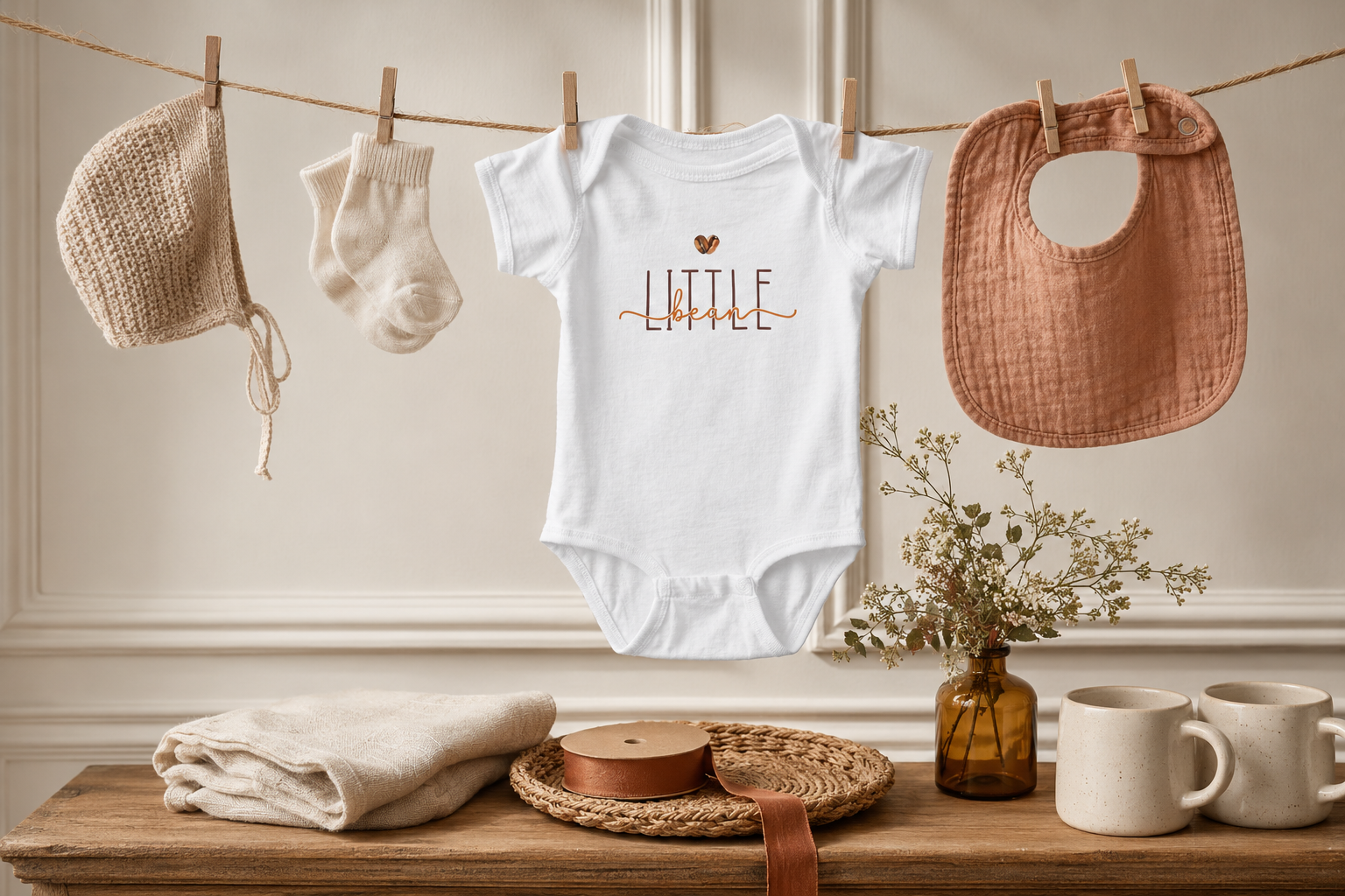 Coffee-themed baby shower clothesline with a “Little Bean” bodysuit, neutral baby accessories, warm wood, linen, amber glass, and greenery.