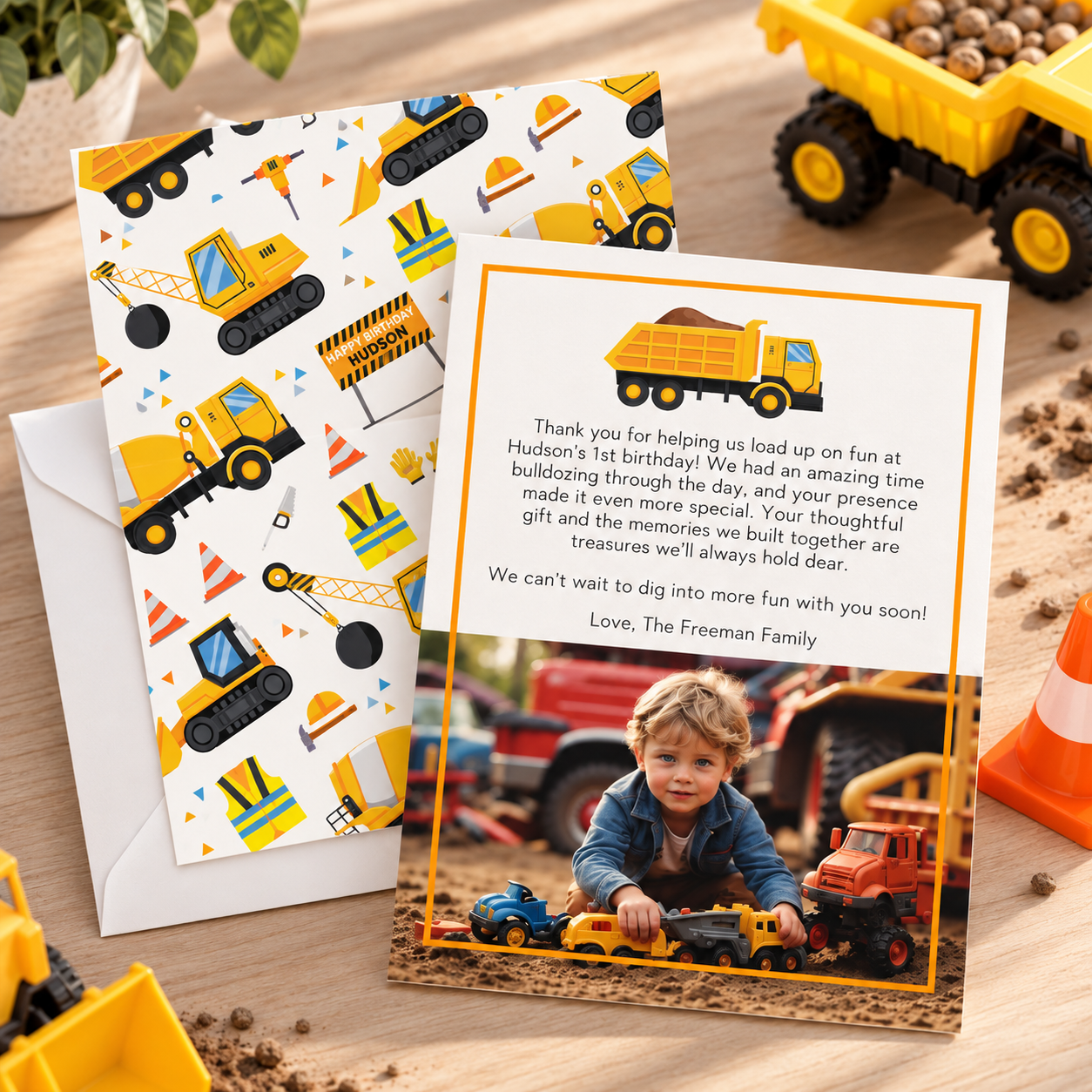 Construction birthday thank-you card with dump truck design, personalized message and toddler photo, styled with matching construction-patterned envelope liner, toy dump trucks, and orange traffic cone.