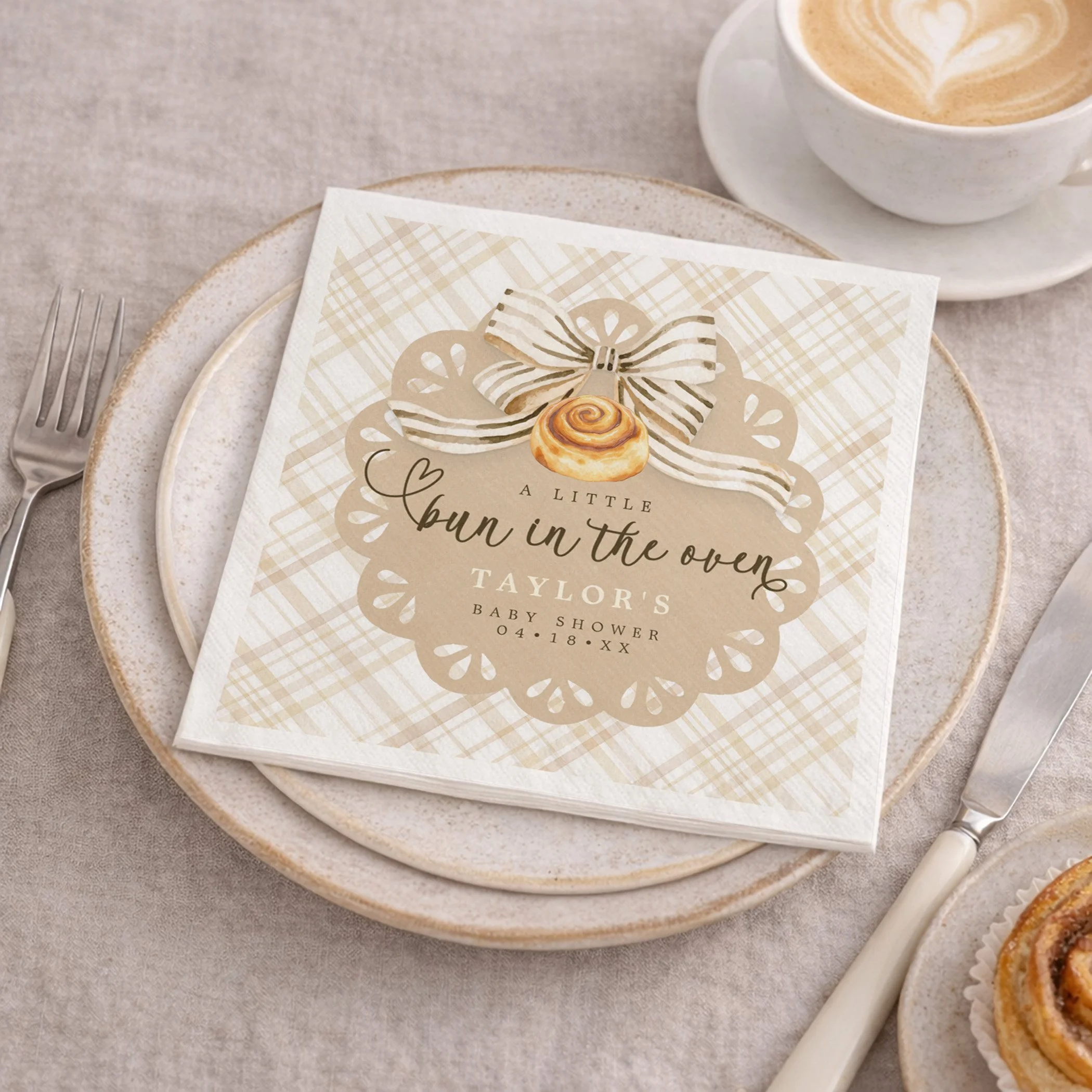 Bun in the Oven baby shower napkins with beige gingham design placed on a neutral table setting with plates, utensils, coffee, and cinnamon roll.
