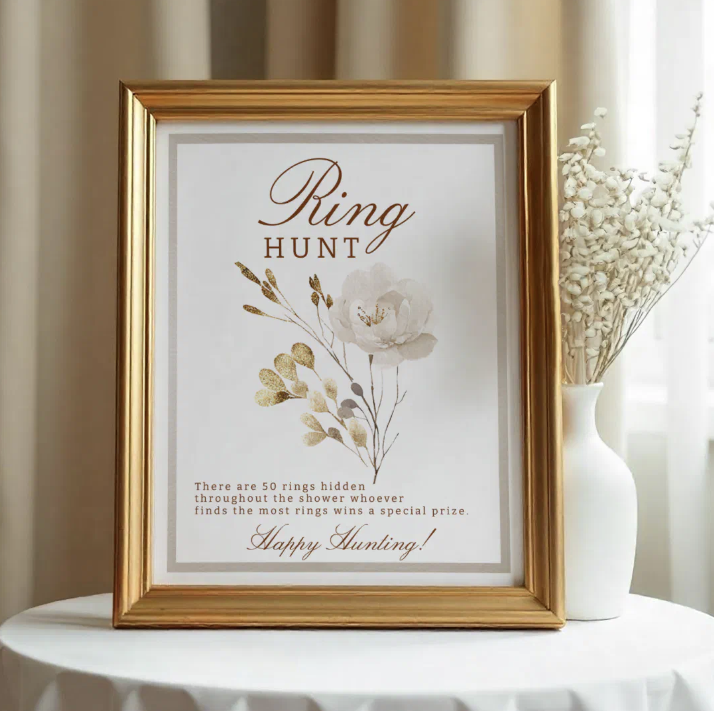 Elegant Ring Hunt bridal shower game sign displayed in a gold frame on a round table beside a white vase with dried florals. Printable bridal shower activity sign with floral design and instructions for hidden ring game.