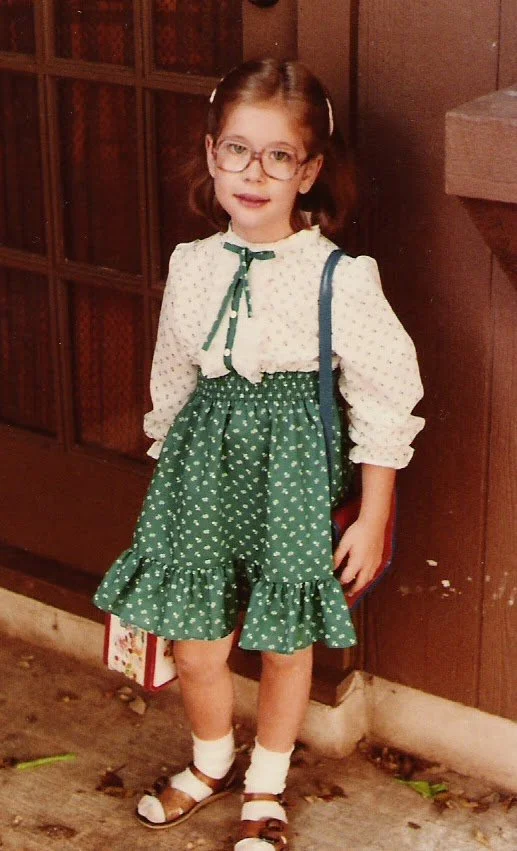 A young girl with glasses and brown hair wearing a white blouse with a green ribbon, a green skirt with white polka dots, white socks, and brown sandals, standing in front of a wooden door.
