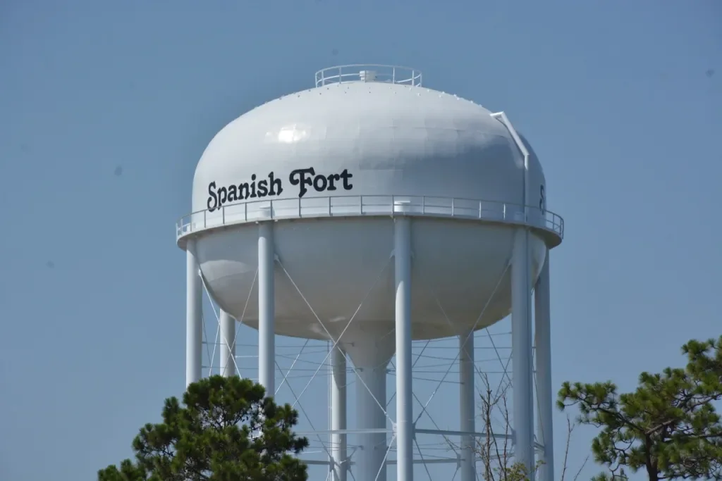 Water tower with the words 'Spanish Fort' written on it, against a blue sky, partially obscured by trees.